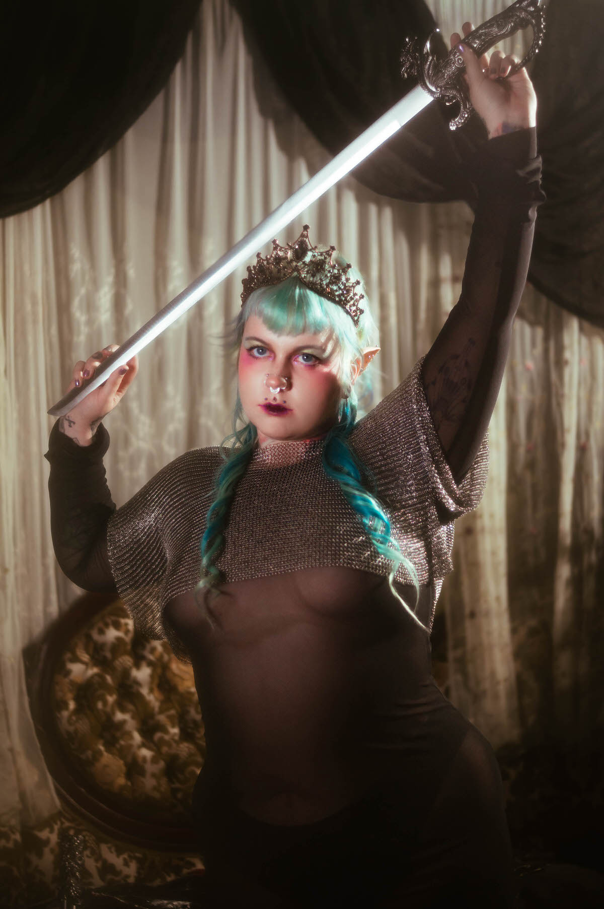 A tattoo artist branding photoshoot image of a woman raising a silver hilted sword overhead, wearing a dark jeweled tiara and a metallic chainmail top over a sheer black mesh dress, lit against striped curtains, Royal Lune Photo.