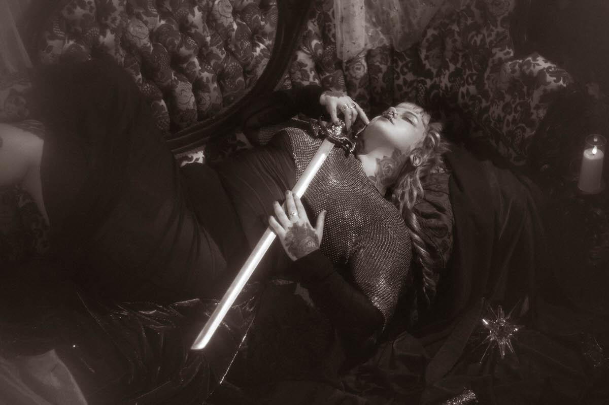 A black and white fantasy branding photoshoot image of a woman reclining with a glowing bladed sword held against her chainmail top, lit by a single candle beside her, Royal Lune Photo