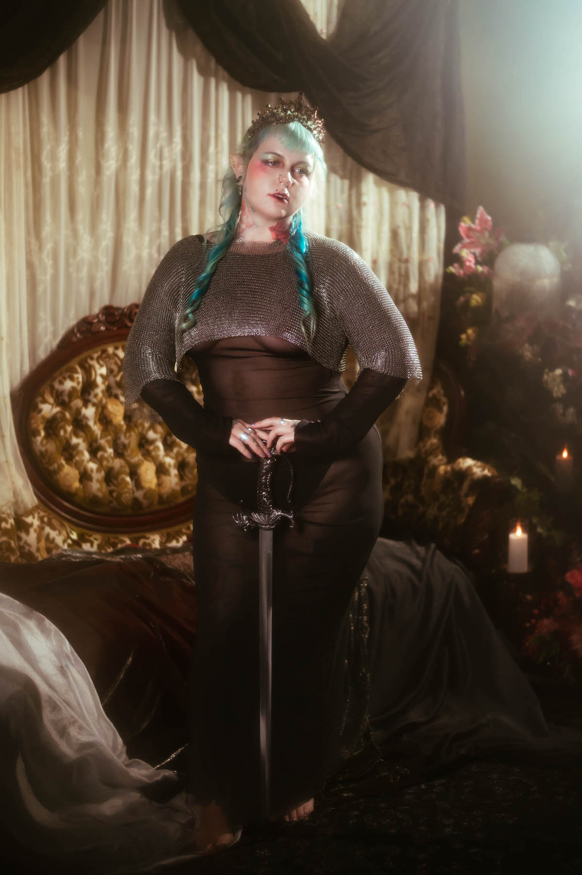 A fantasy branding photoshoot portrait of a woman standing with hands on her hips, a sword resting point down in front of her, wearing a chainmail top, black mesh dress, and dark tiara beside lit candles, Royal Lune Photo.