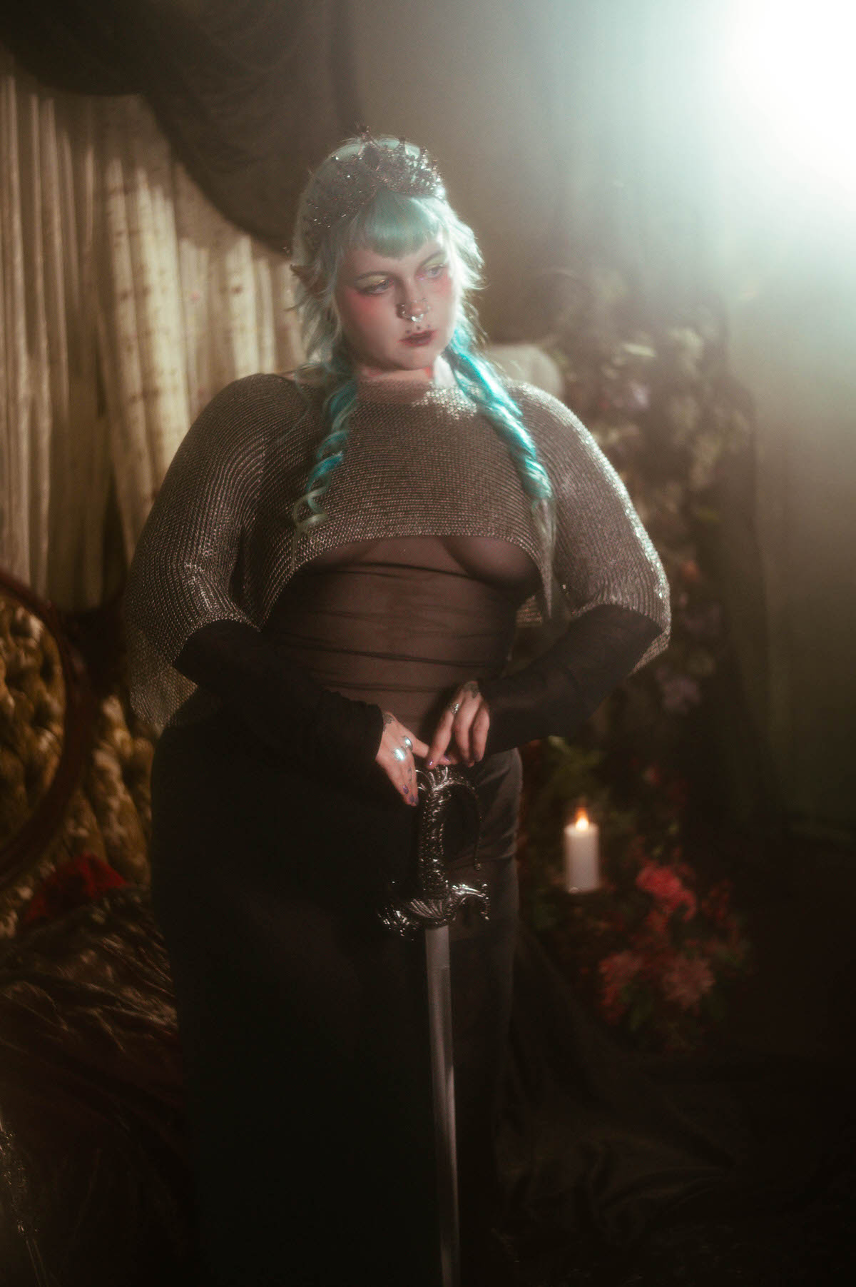 A tattoo artist branding photography image of a woman seated in an ornate wooden chair, resting a silver hilted sword against her shoulder, wearing a chainmail top and dark tiara beside a lit candle, Royal Lune Photo.