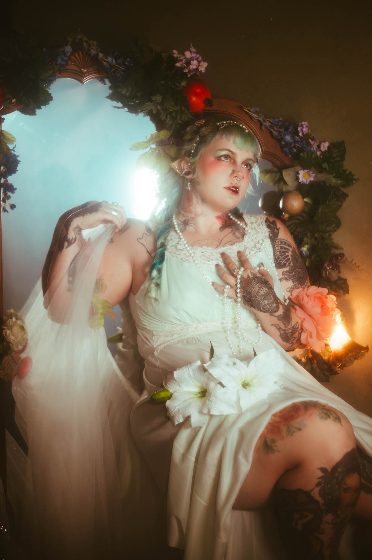 A tattoo artist branding photoshoot portrait of a woman holding sheer tulle fabric and white lily flowers while seated beside a vanity mirror, pearls draped over her chest, lit by warm candlelight, Royal Lune Photo.