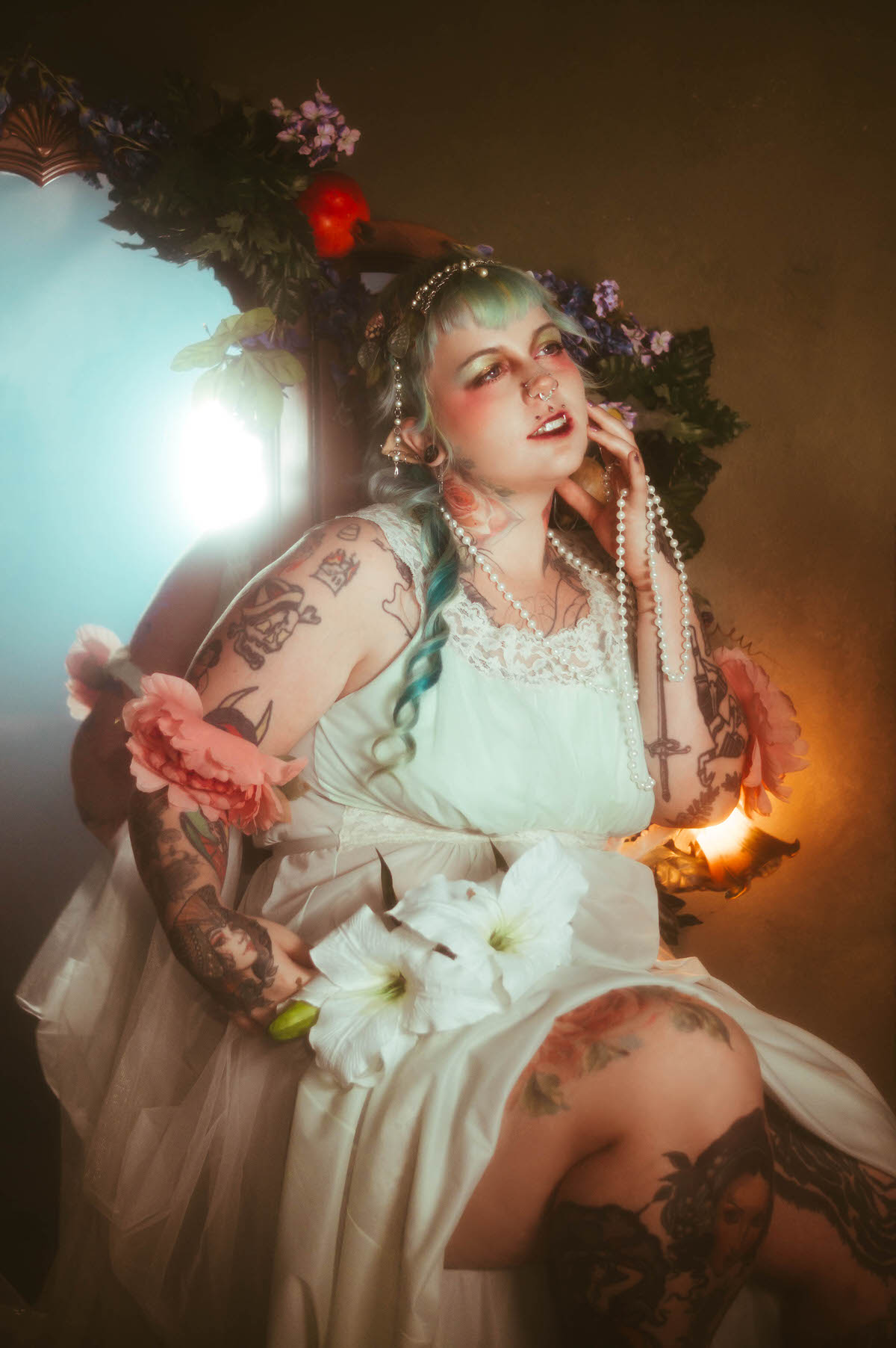 A tattoo artist branding photoshoot portrait of a woman with mint colored hair holding white lily flowers and a strand of pearls, seated beside a vintage mirror decorated with a floral garland and red apples, softly lit by a warm golden lamp at Royal Lune Studio in Dallas, TX, Royal Lune Photo.