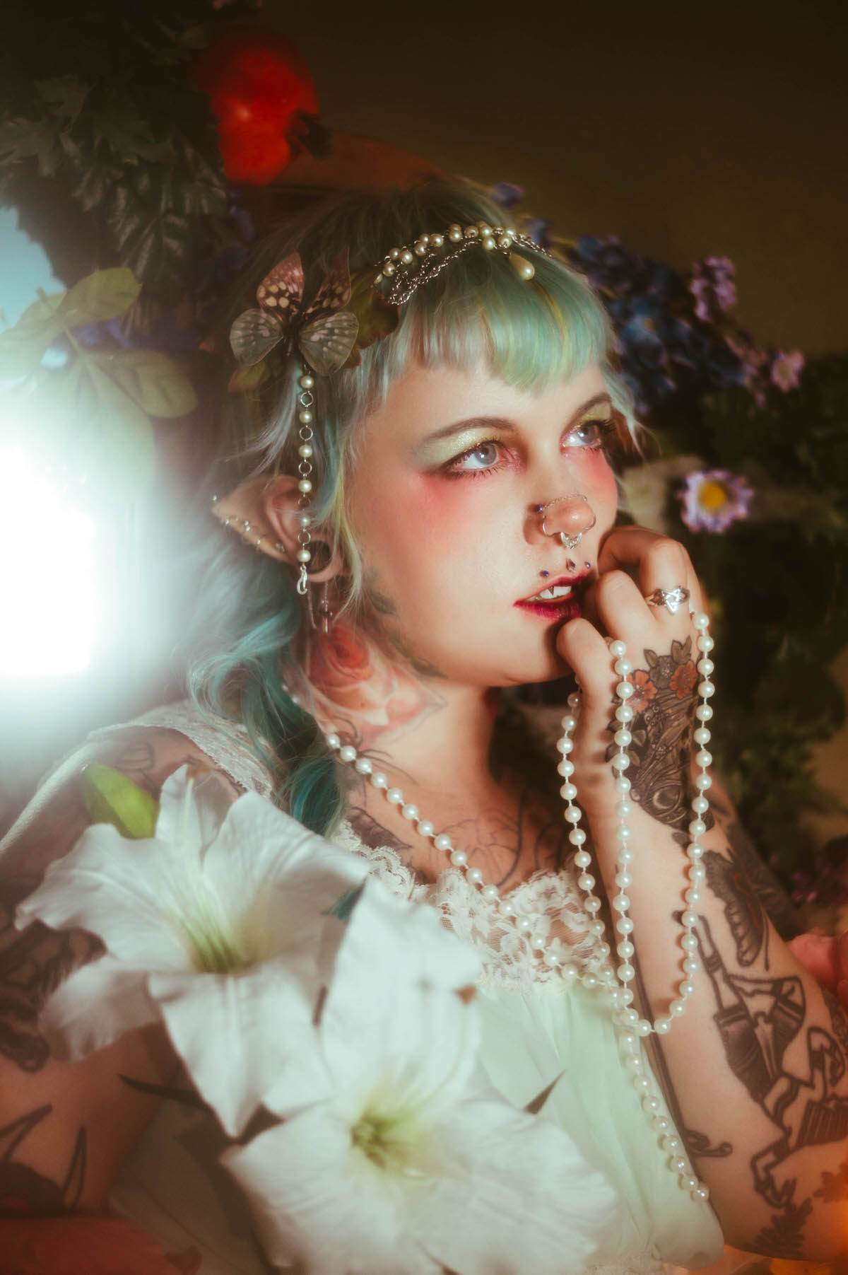A close up content photography for creatives in Dallas portrait of a woman with a pearl and butterfly headpiece, looking upward while holding pearls near her face beside white lily and pink peony flowers, Royal Lune Photo.