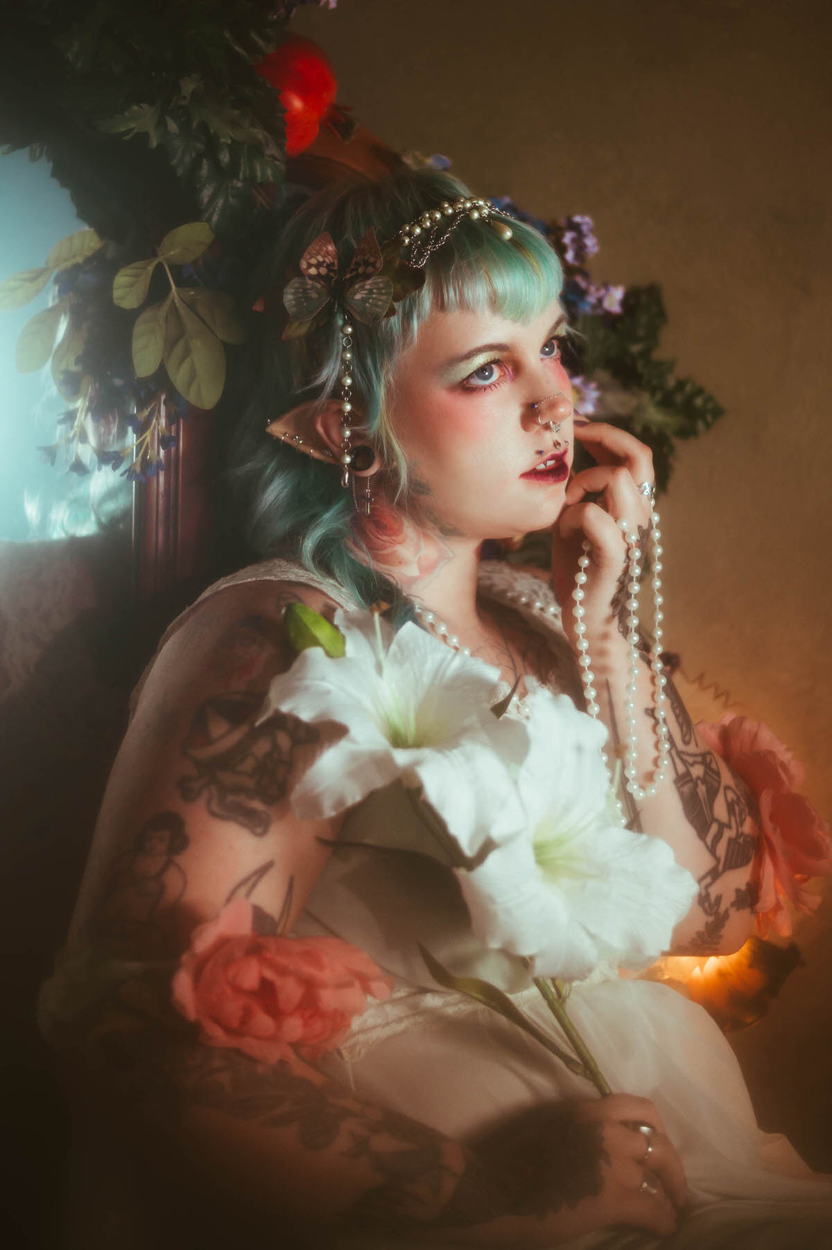 A fantasy branding photoshoot close up portrait of a woman looking upward while holding a strand of pearls near her chin, her septum piercing and fangs visible beside a white lily flower, Royal Lune Photo.