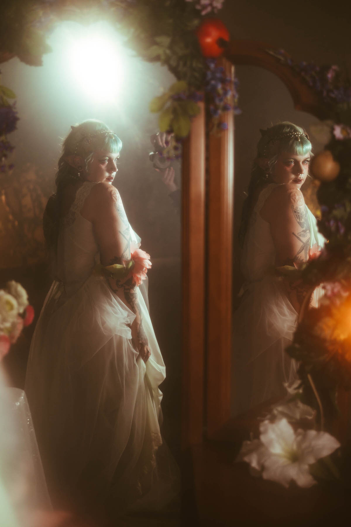A tattoo artist branding photoshoot double exposure image showing a woman in a pale mint nightgown reflected beside an antique mirror decorated with a floral garland and red apples, her tattooed back visible under soft blue and warm lighting, Royal Lune Photo.