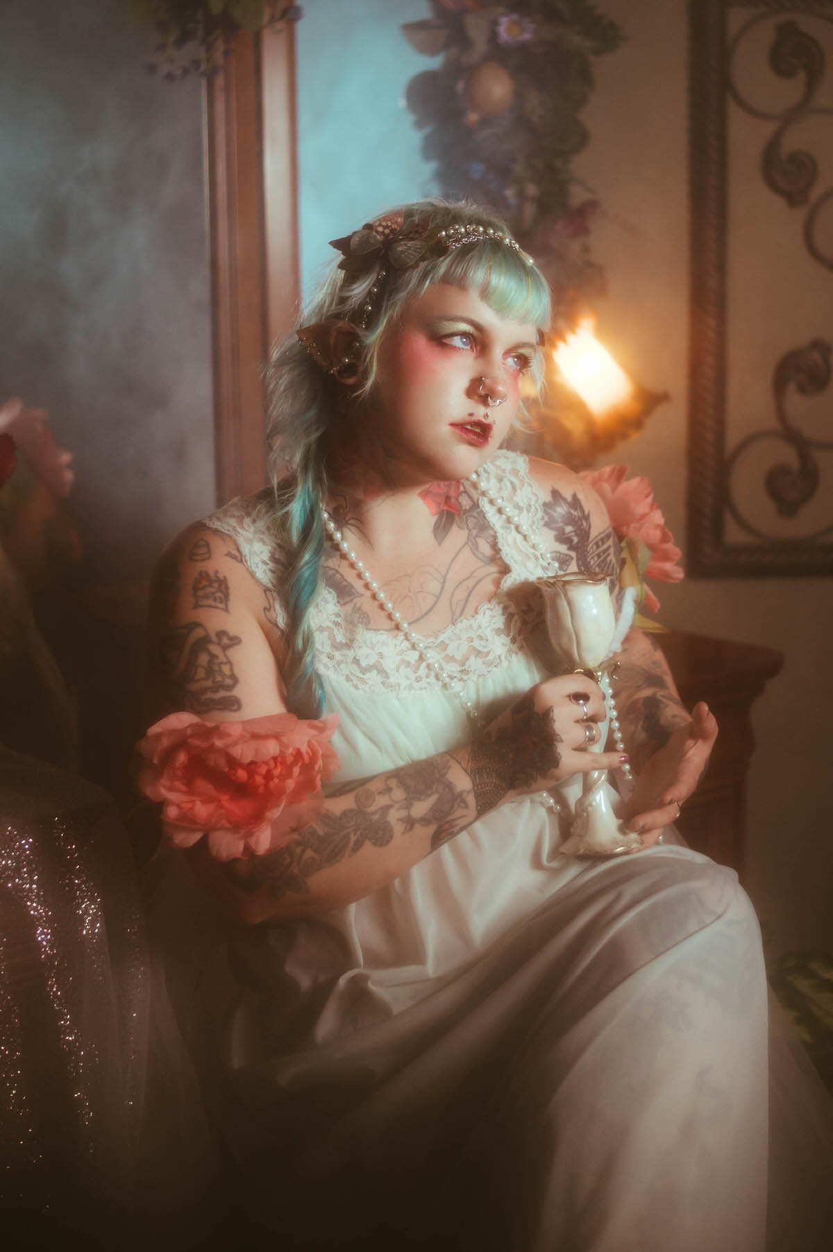 A personal branding photos for artists image of a woman seated holding an ornate white chalice and a pink peony flower, pearls draped across her tattooed arm in front of a vanity mirror, Royal Lune Photo.