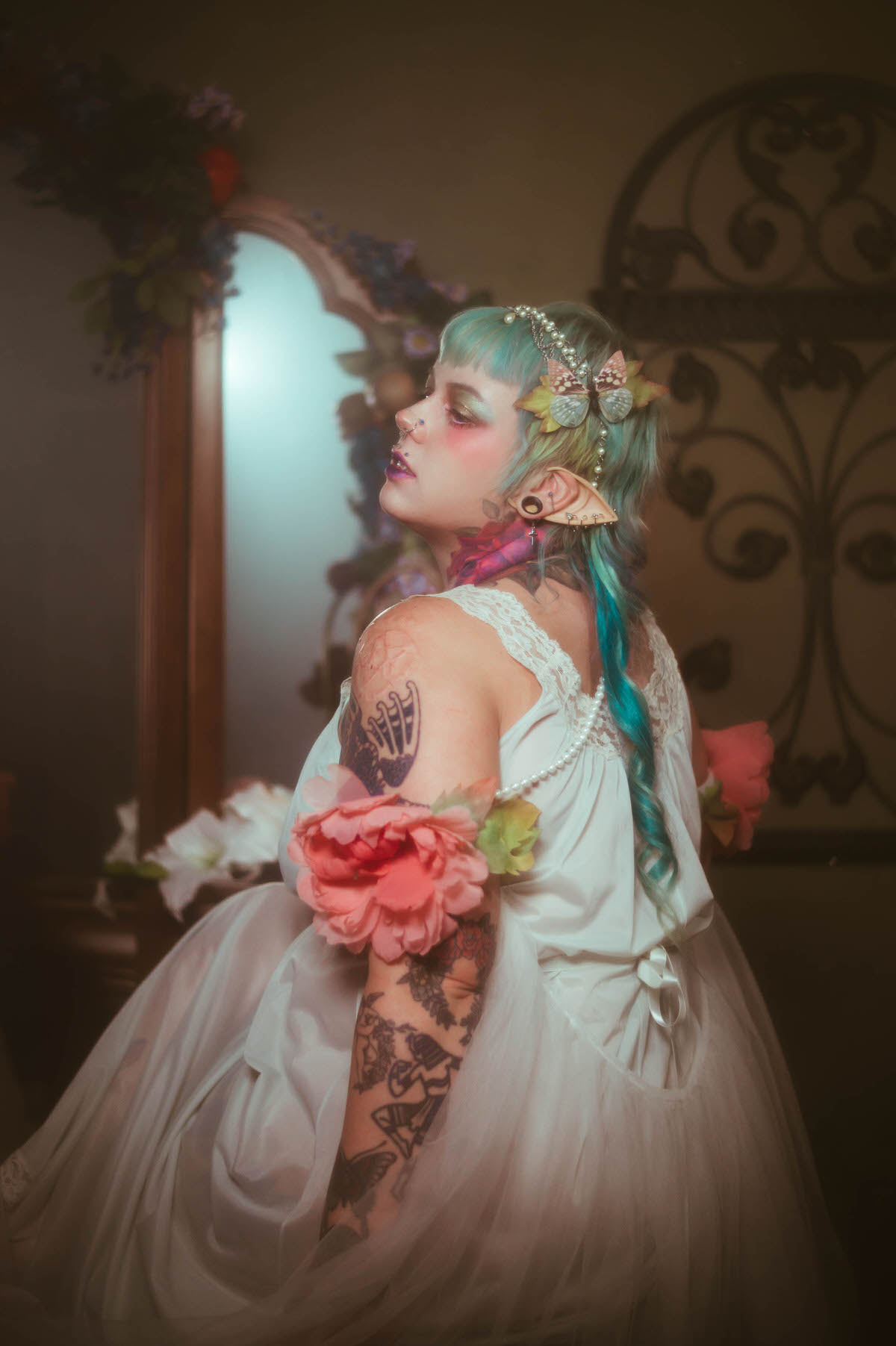 A tattoo artist branding photography side profile portrait of a woman with a pink peony and butterfly clip in her hair, her tattooed back visible beside an ornate wrought iron wall decoration, Royal Lune Photo.