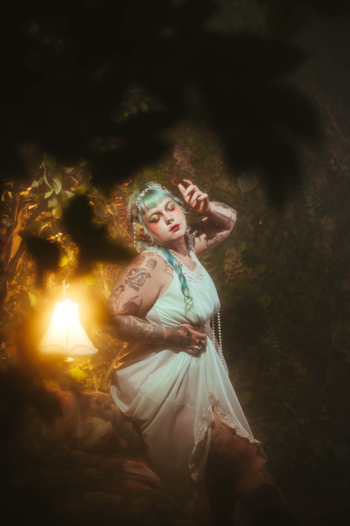 A content photography for creatives in Dallas portrait of a woman standing among greenery with one hand raised to her head, eyes closed, her tattooed arm visible in warm lamp light, Royal Lune Photo.