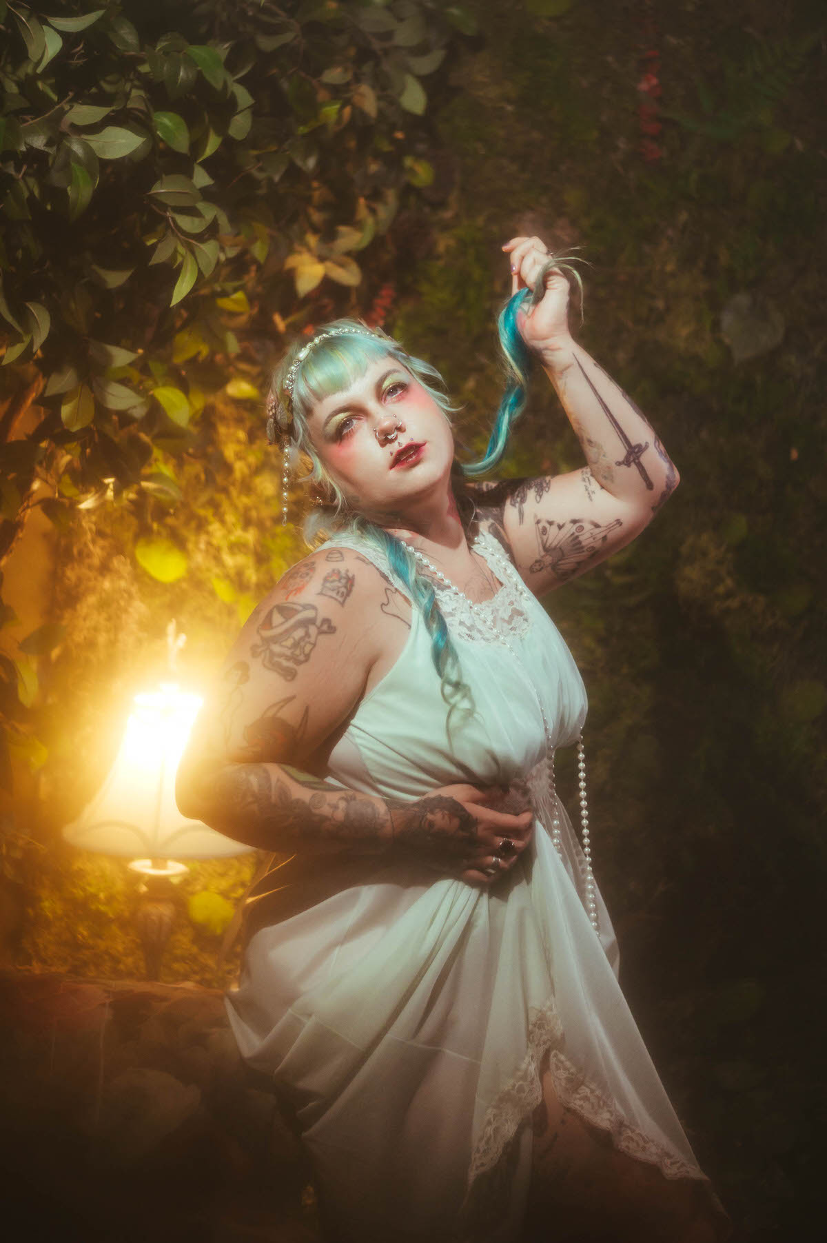 A tattoo artist branding photoshoot image of a woman standing in a garden setting, holding a strand of her teal hair upward with her tattooed arm raised, pearls draped over her pale dress in warm golden lamplight, Royal Lune Photo.