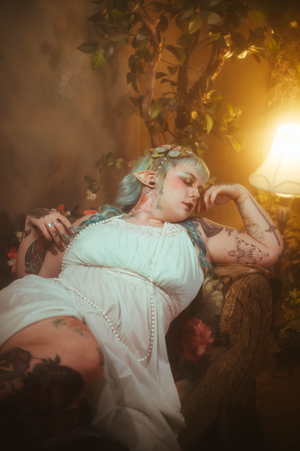 A tattoo artist branding photoshoot image of a woman reclining on a vintage sofa in a mint green nightgown, resting her head on her tattooed arm beside a warm lamp and climbing greenery, Royal Lune Photo.