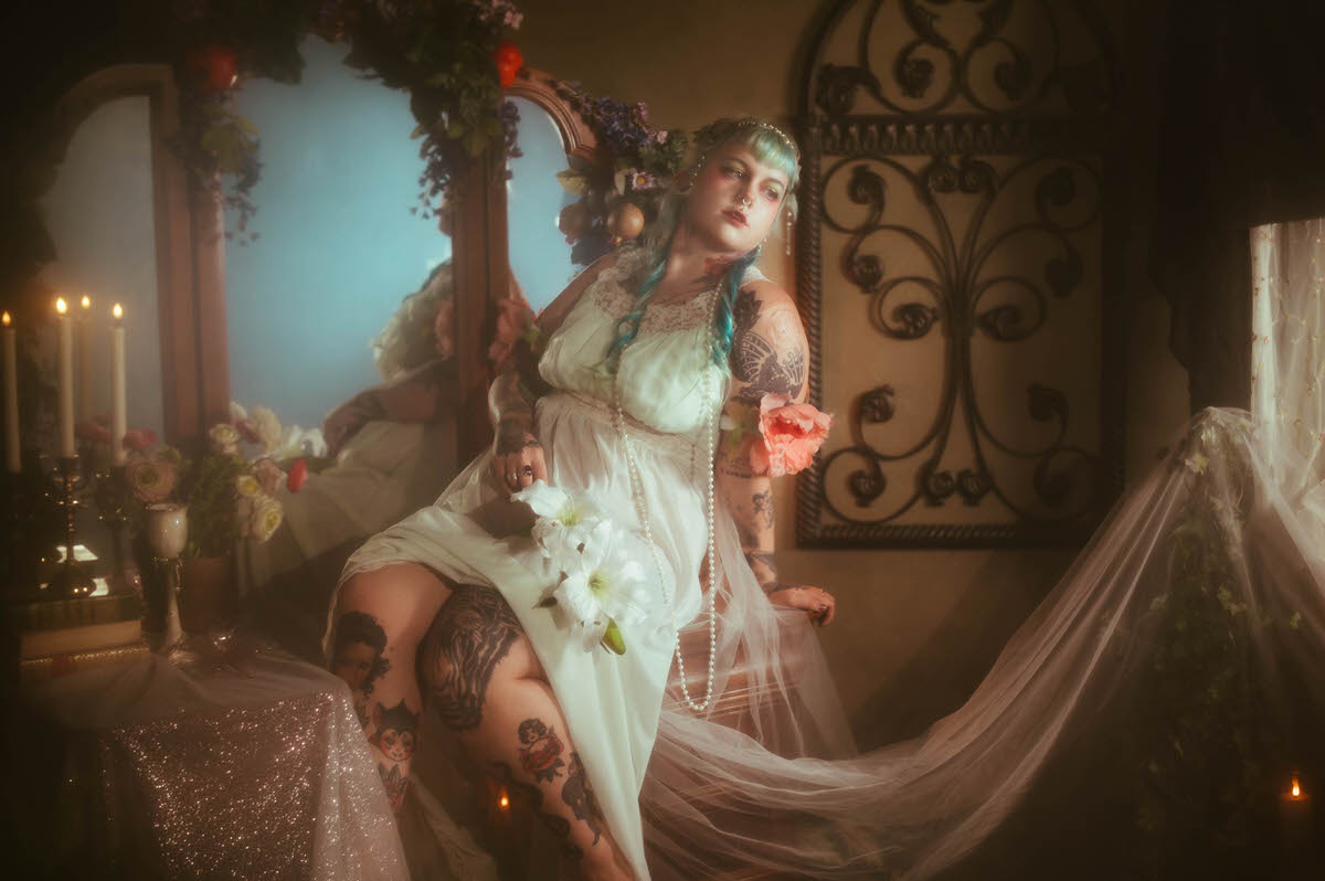 A tattoo artist branding photography image of a woman seated beside a vanity mirror framed with a floral garland, holding a white lily flower and a strand of pearls with her tattooed legs visible beneath her pale mint dress at Royal Lune Studio, Royal Lune Photo.