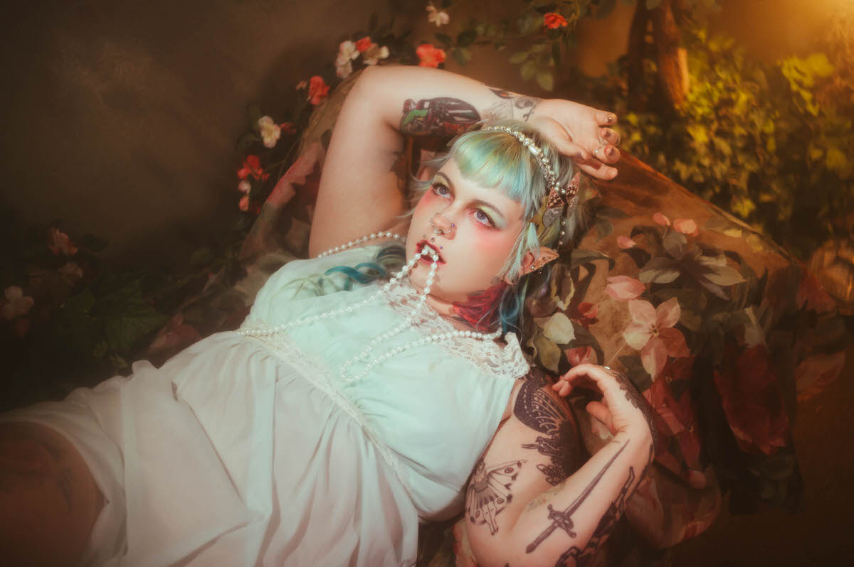 A fantasy branding photoshoot overhead portrait of a woman lying among floral fabric in a mint green nightgown, a strand of pearls draped across her lips, surrounded by soft red and white flowers, Royal Lune Photo.