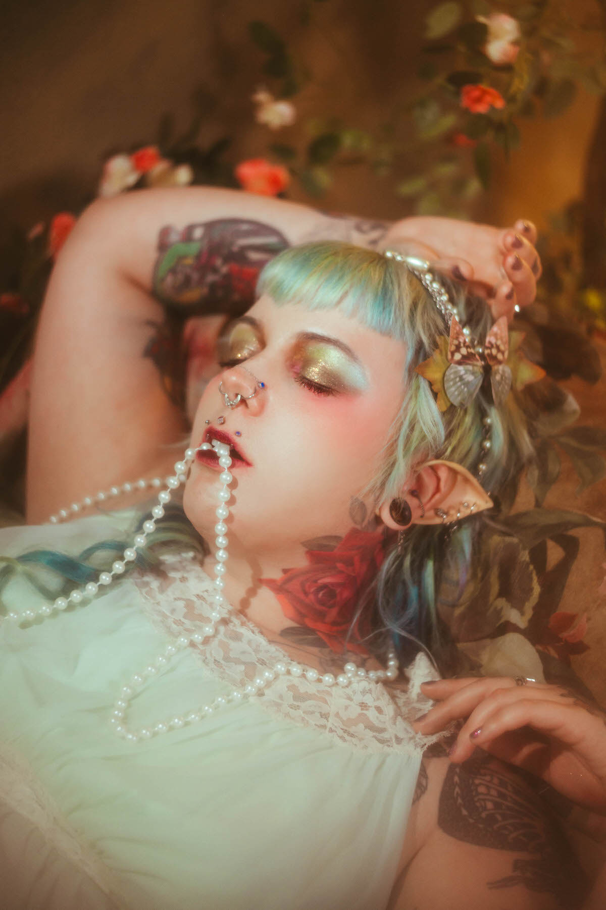 A close up tattoo artist branding photography image of a woman lying back with her eyes closed, gold and pink shimmer eye makeup, a strand of pearls held between her lips, and a red rose tattoo visible on her neck, Royal Lune Photo.