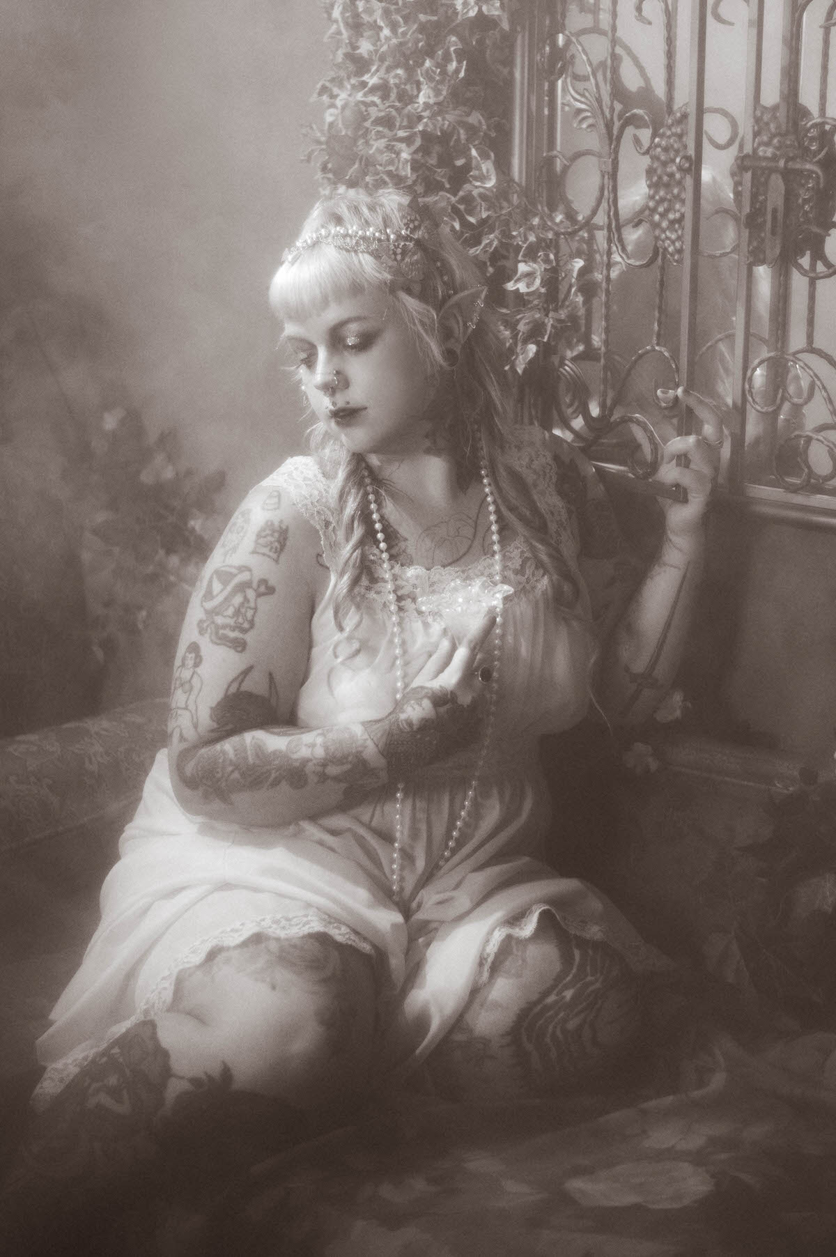 A black and white tattoo artist branding photoshoot portrait of a woman seated with her tattooed arm resting across her knee, holding a cigarette beside an ornate wrought iron gate, Royal Lune Photo.
