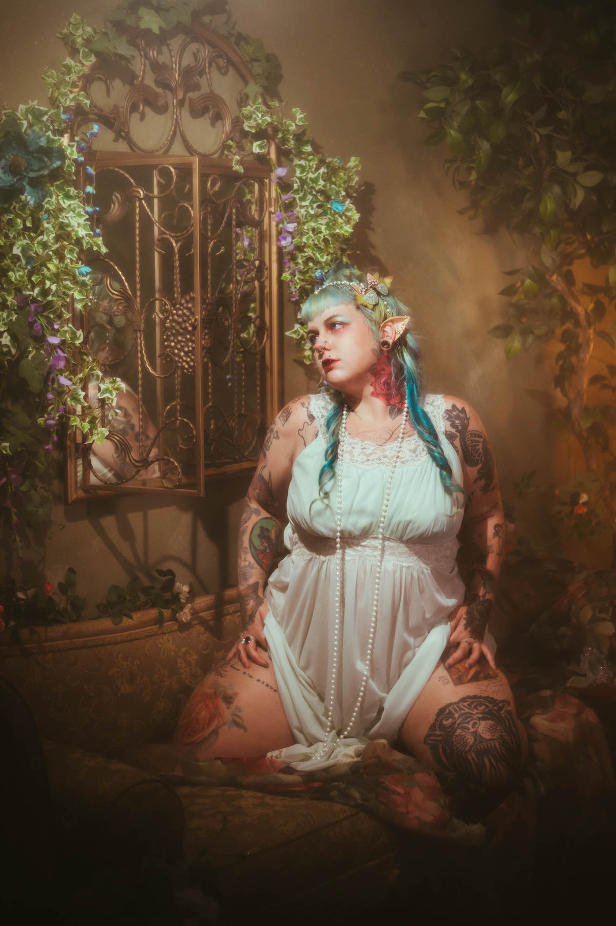 A personal branding photos for artists image of a woman kneeling in a mint green nightgown beside an ornate gold framed mirror decorated with ivy and purple flowers, her tattooed legs and arms visible in warm lamplight, Royal Lune Photo.