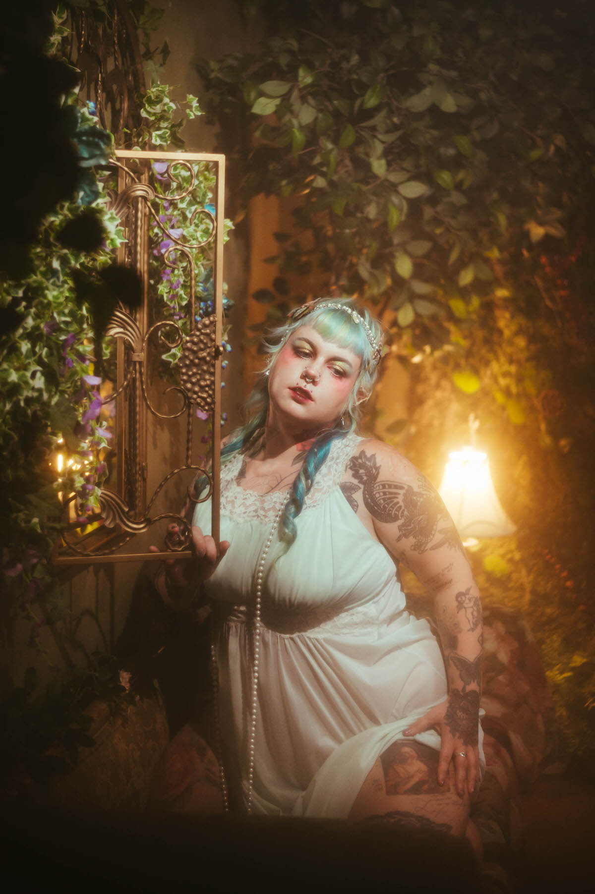 A Dallas Fort Worth branding photographer image of a woman in a mint green nightgown standing beside a gold framed mirror surrounded by climbing ivy and purple flowers, her tattooed arm resting on the frame, Royal Lune Photo.