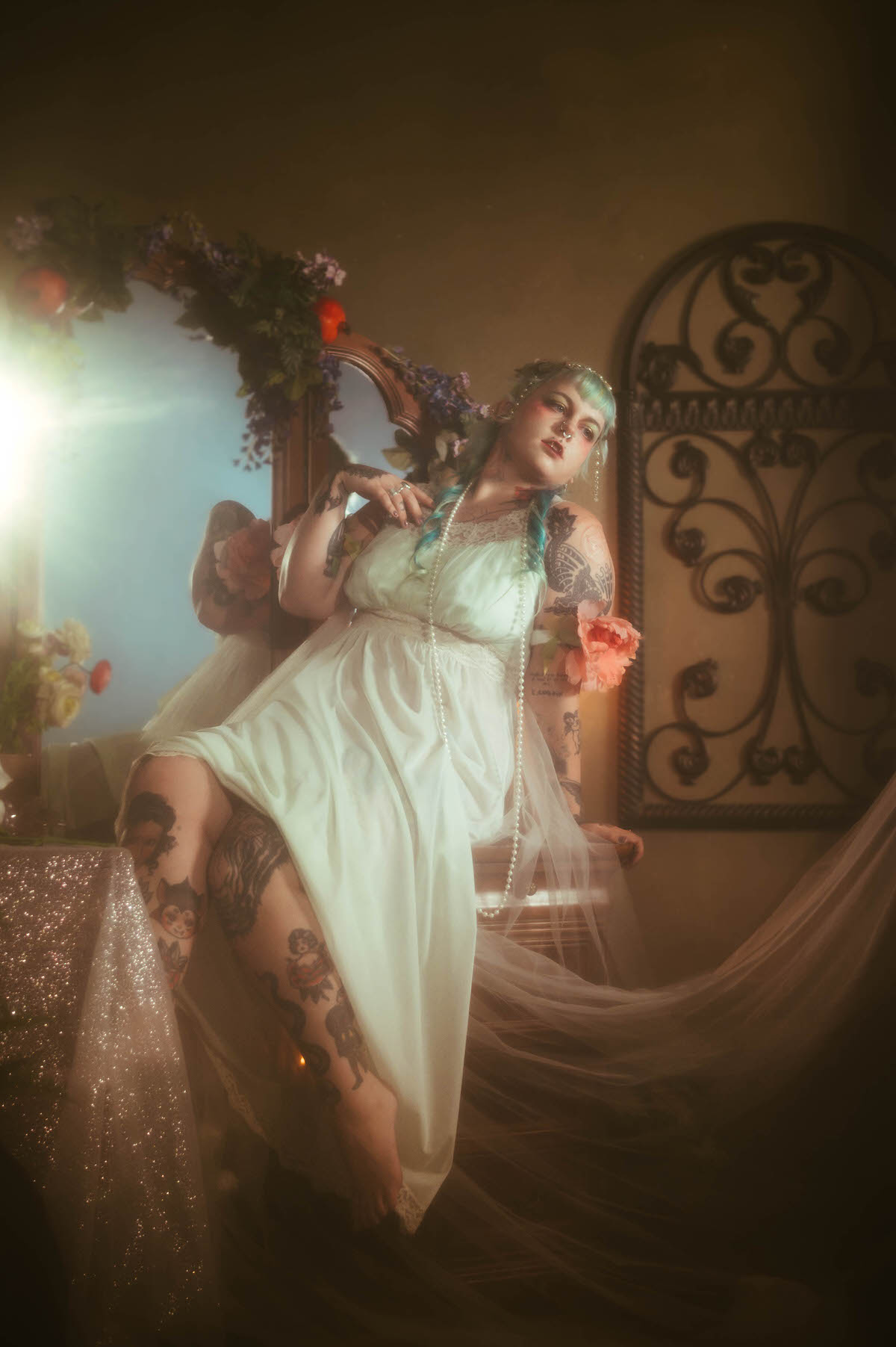 A personal branding photos for artists image of a woman seated with one tattooed leg raised, holding a pink peony and pearls, in front of a vanity mirror and ornate wrought iron wall art, Royal Lune Photo.