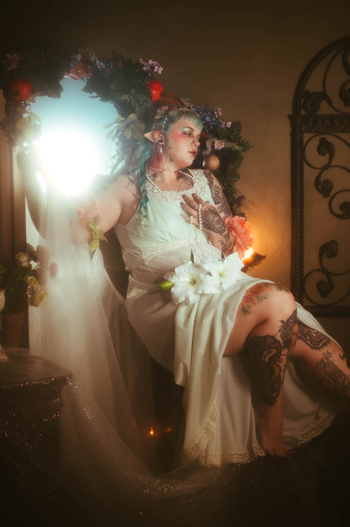 A Dallas Fort Worth branding photographer image of a woman reclining with white lily flowers in her lap and pearls draped across her chest, her tattooed leg extended beside a floral framed mirror, Royal Lune Photo.