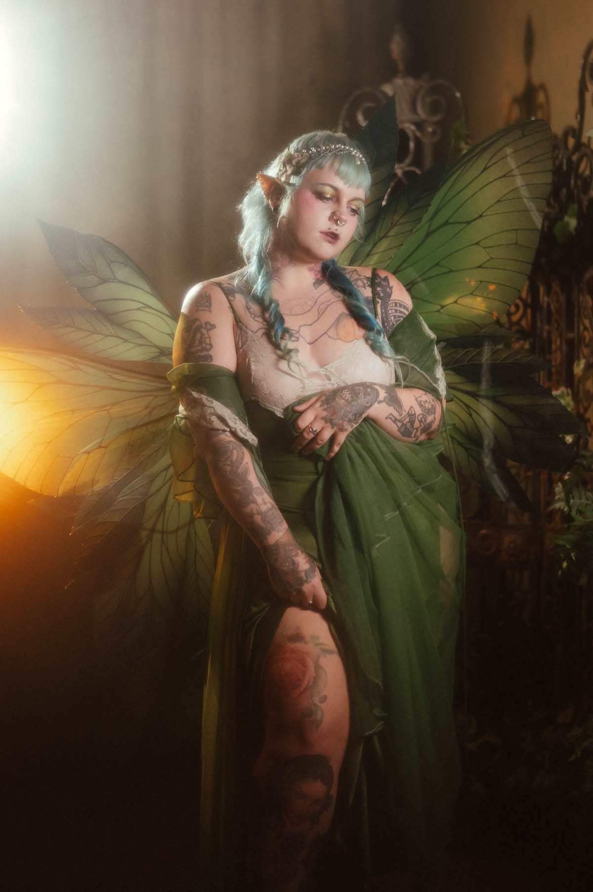 A fantasy branding photoshoot image of a woman in a green fairy dress and prosthetic elf ears standing beside an ornate wrought iron gate, looking down at her tattooed leg with a rose tattoo visible through the sheer fabric, Royal Lune Photo.