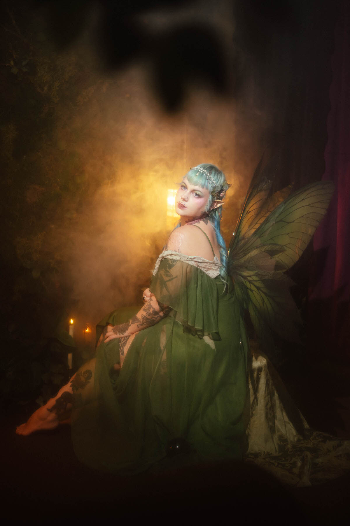 A Dallas Fort Worth branding photographer image of a woman looking back over her shoulder in a sage green off the shoulder dress with lace sleeve trim and large green fairy wings, lit by warm candlelight and soft haze, Royal Lune Photo.