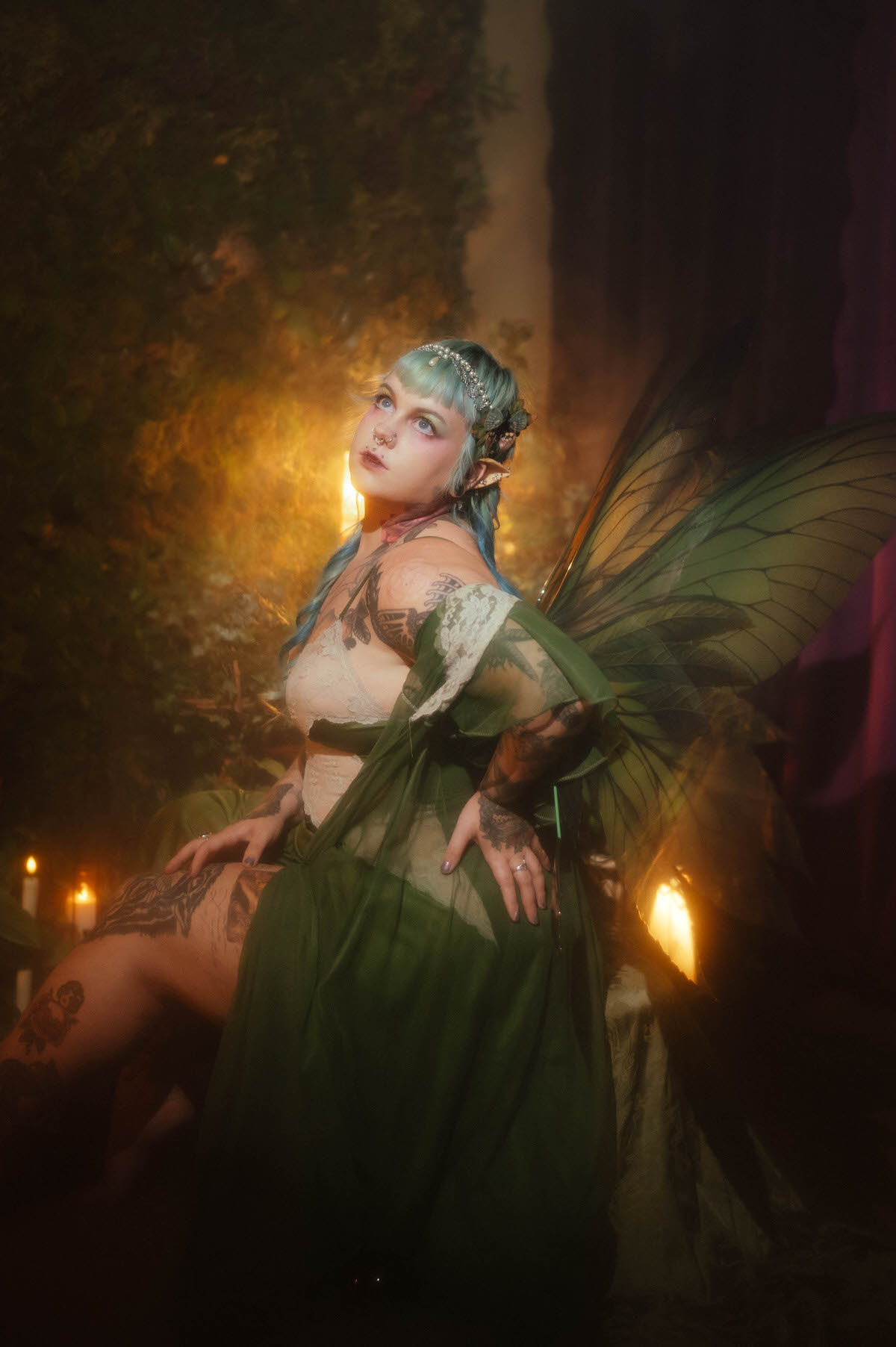 A tattoo artist branding photoshoot portrait of a woman standing in a sage green dress with sheer lace sleeves and fairy wings, gazing upward beside lit candles with her tattooed thigh visible through the fabric, Royal Lune Photo.