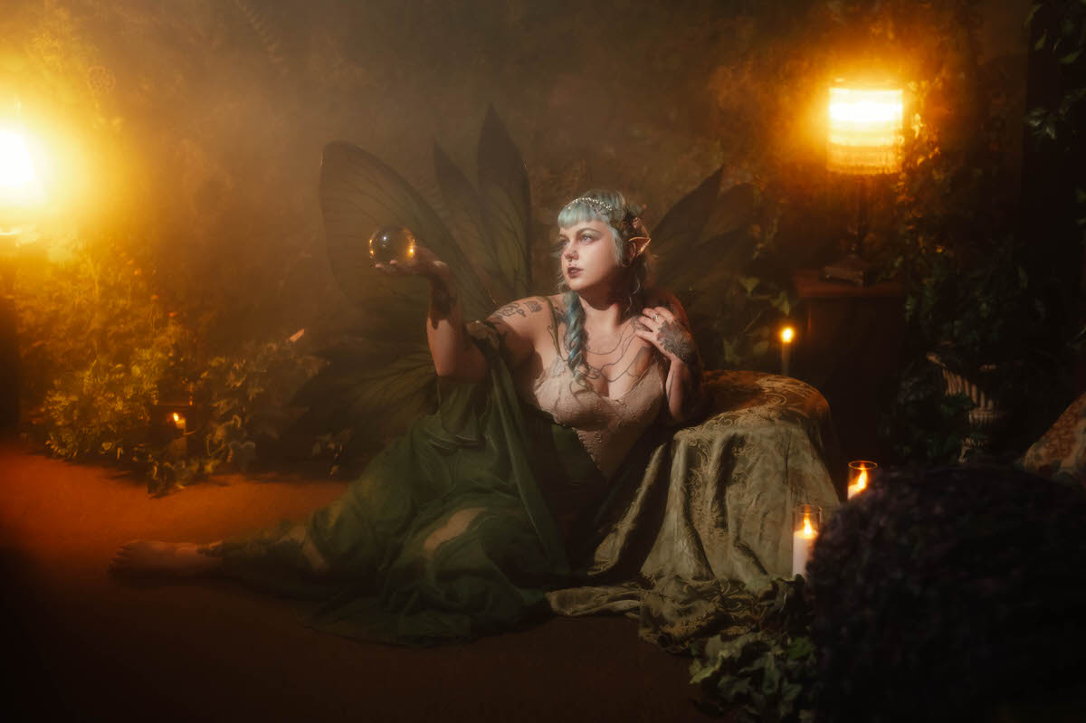 A tattoo artist branding photography image of a woman reclining on a chaise in a sage green dress with fairy wings from Wings and Flower Design, holding a glowing amber orb while surrounded by warm lantern light and lit candles at Royal Lune Studio, Royal Lune Photo.