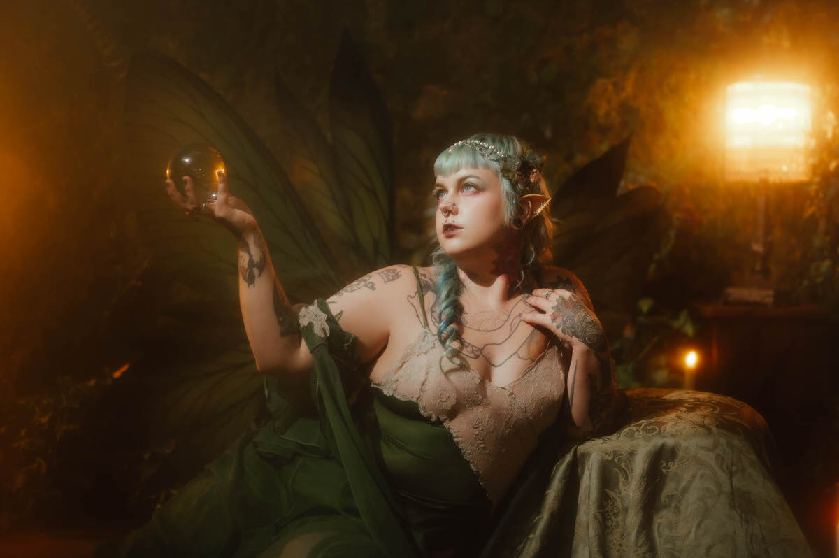 A content photography for creatives in Dallas portrait of a woman with septum and philtrum piercings, a tattooed chest and arms, and green fairy wings, gazing to the side while holding an amber glass orb near warm lantern light, Royal Lune Photo.