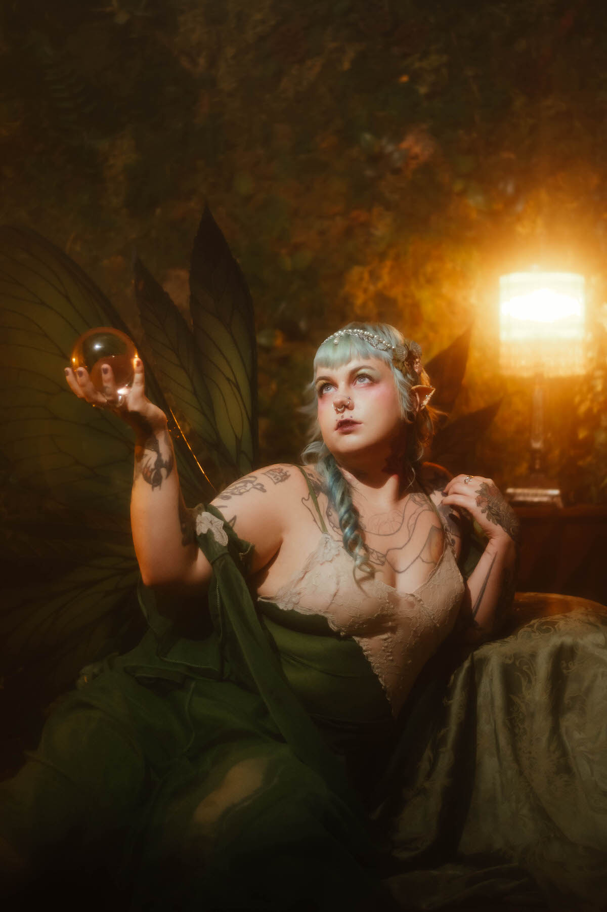 A close up portrait for a personal branding photos for artists session showing a woman with mint blue hair, a pearl and butterfly headpiece, and prosthetic elf ears, holding a glowing glass orb above her tattooed arm in warm golden light, Royal Lune Photo.