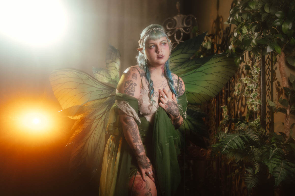 A tattoo artist branding photography portrait of a woman with fairy wings and elf ears standing among greenery and an ornate wrought iron gate, one hand resting on her chest, softly lit by warm backlighting at Royal Lune Studio, Royal Lune Photo.