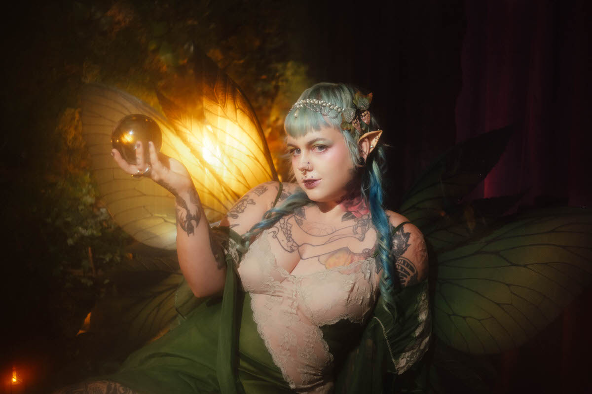A fantasy branding photoshoot image of a woman wearing Wings and Flower Design fairy wings and prosthetic elf ears while holding a dark glass orb, styled with an ivory lace bodice and a pearl headpiece with a butterfly clip, glowing in warm golden lantern light at Royal Lune Studio, Royal Lune Photo.