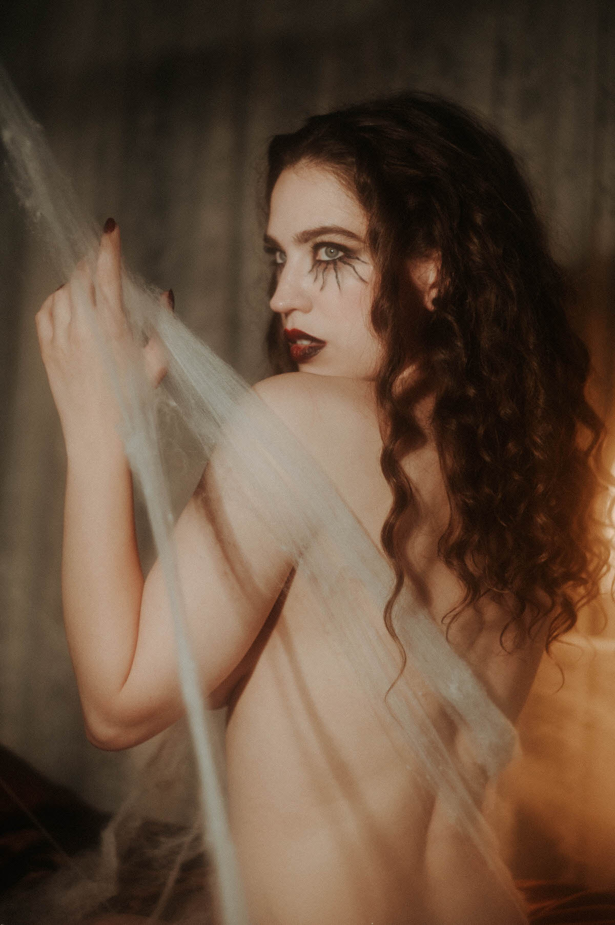 Photographed from the side, a woman reaches up to hold sheer grey cobweb netting, her bare back lit with warm amber tones on one side and cool shadow on the other, showing off her spider leg lashes and dark red nails for this dark fantasy photography session at Royal Lune Photo.