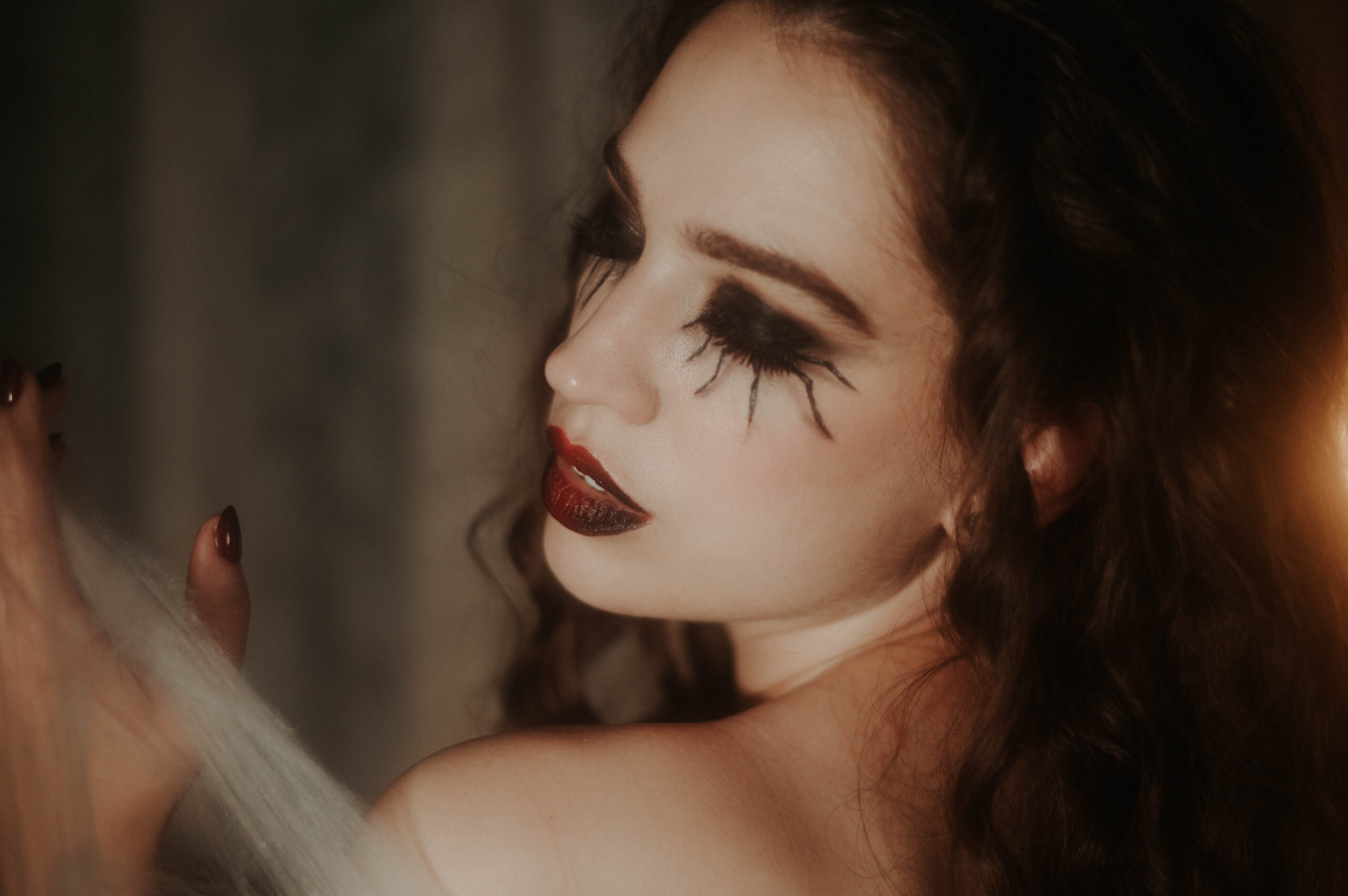 A close up profile portrait captures a woman's dramatic spider leg lashes and deep red lipstick as she reaches toward pale cobweb netting under soft warm lighting for this sfx makeup photoshoot at Royal Lune Photo.