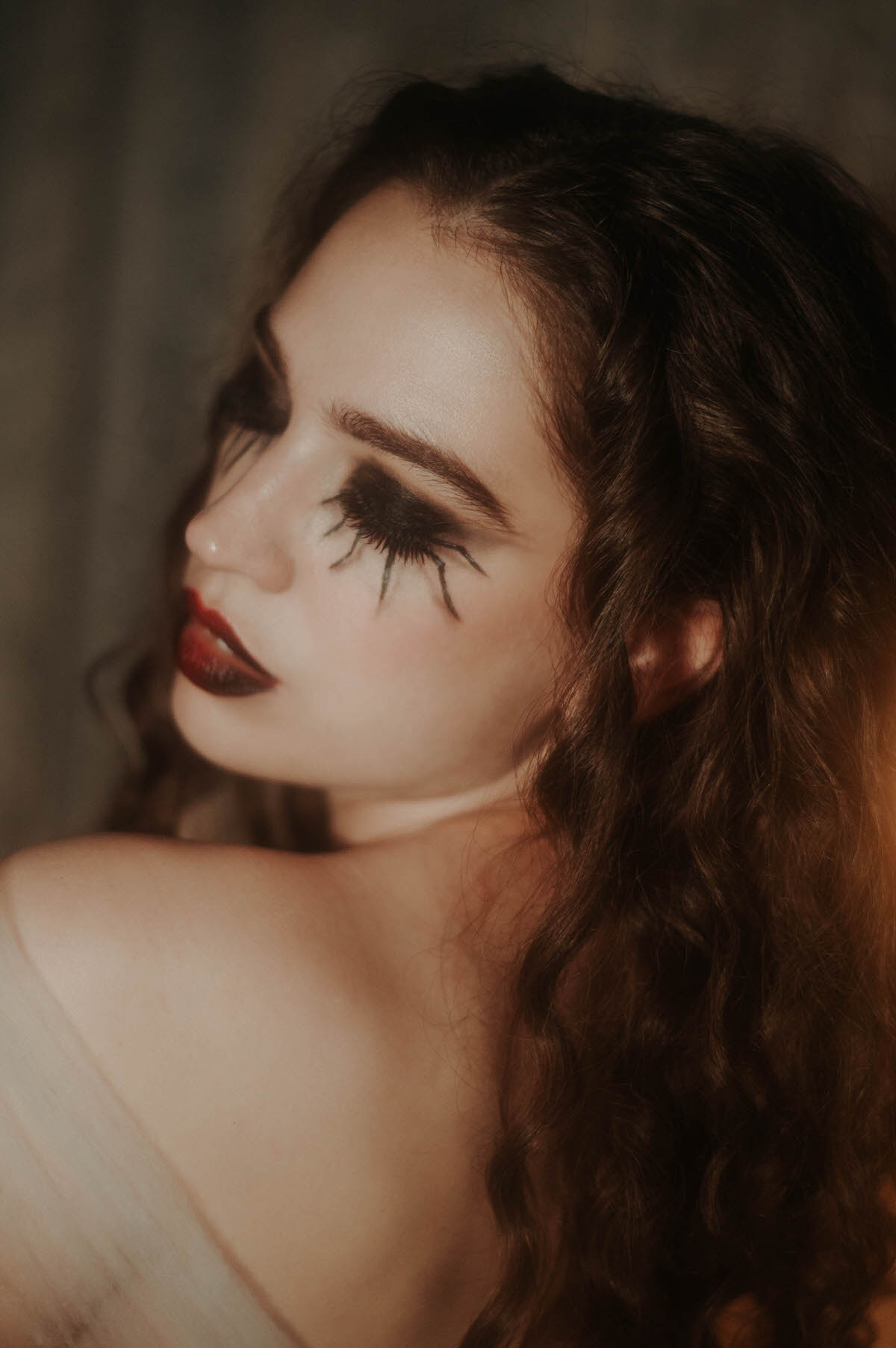 A moody profile portrait features a woman with spider leg lash eye makeup and deep red lipstick, her bare shoulder and dark curls glowing in warm amber light against a shadowed backdrop for this gothic glamour photoshoot at Royal Lune Photo.