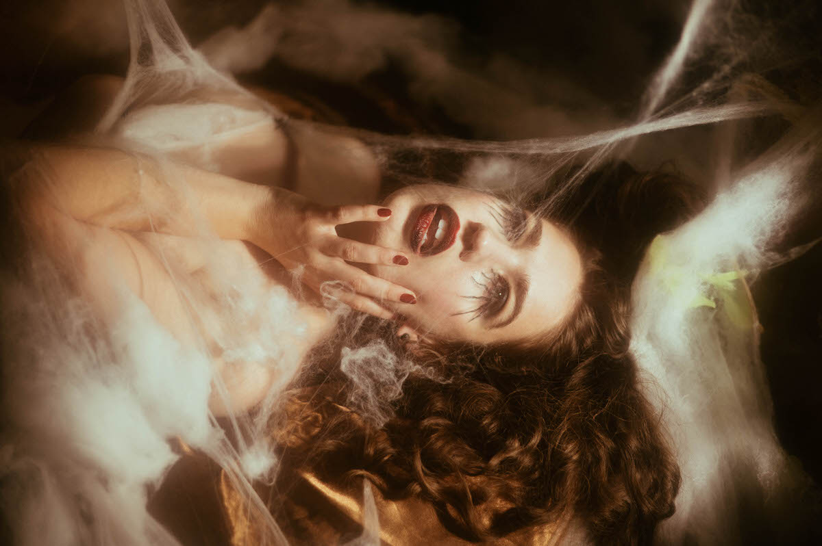 A woman reclines with her hand framing her face, deep red nails and lipstick standing out beneath wispy cobweb netting draped over gold satin sheets for this gothic boudoir photoshoot at Royal Lune Photo.