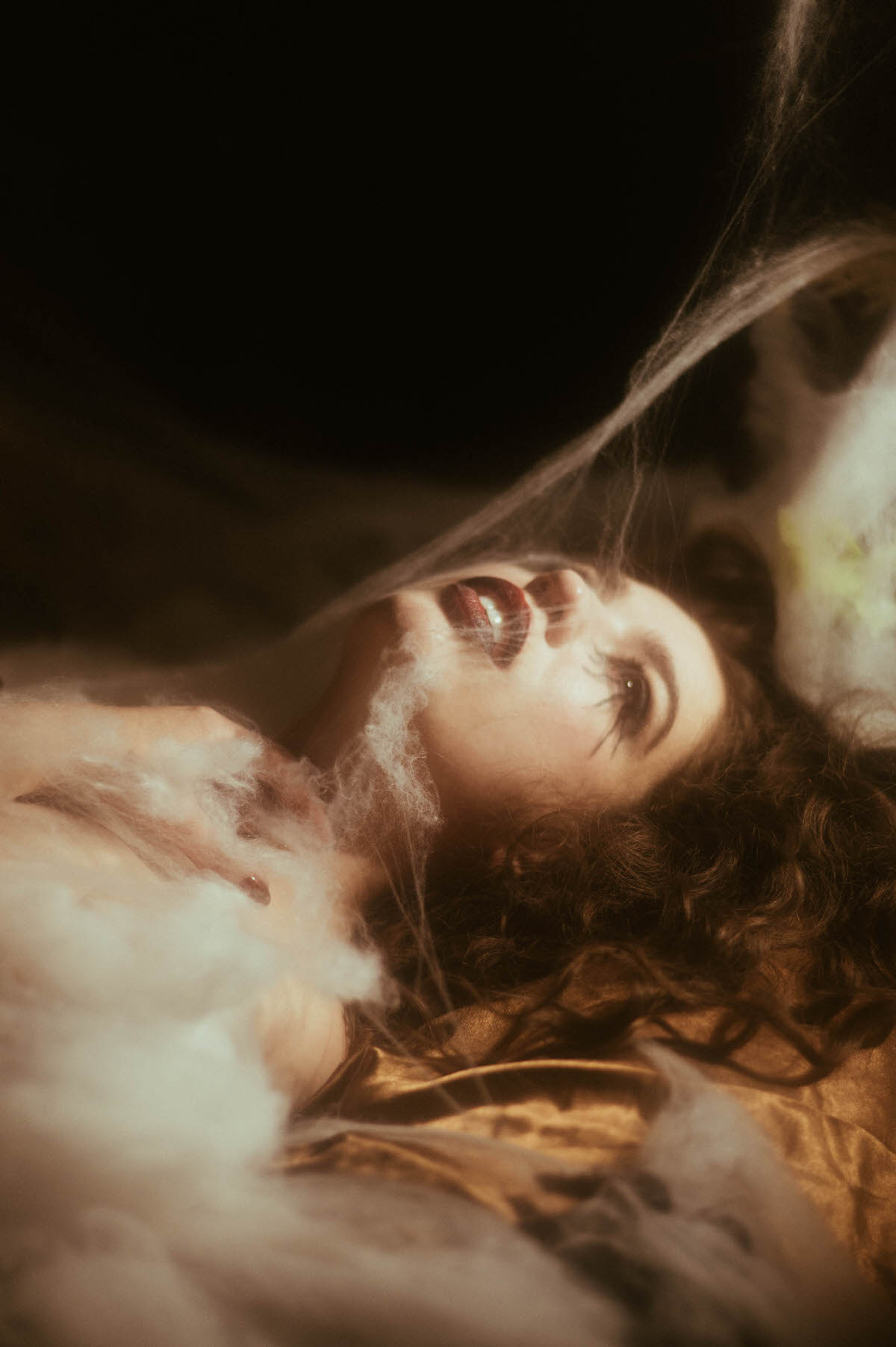 Lying back with her head tilted, a woman reveals deep red lipstick and spider leg lashes through soft cobweb netting draped over gold satin sheets for this dark fantasy photography session at Royal Lune Photo.