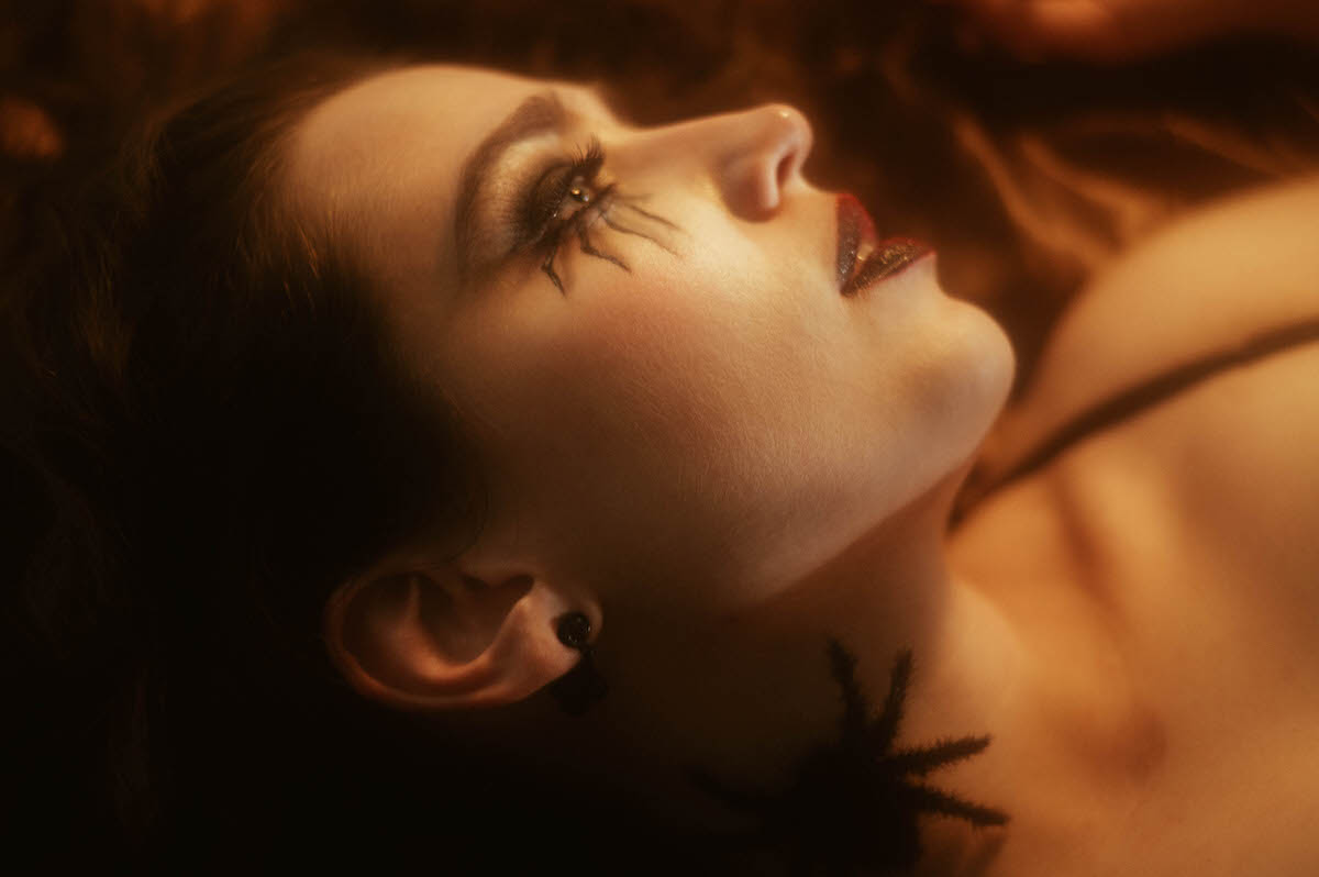 Photographed in close up under warm amber lighting, a woman tilts her head back to reveal dramatic spider leg lash makeup, glossy dark red lipstick, and a black drop earring, with a small prop spider resting against her collarbone atop gold satin sheets for this gothic glamour photoshoot at Royal Lune Photo.