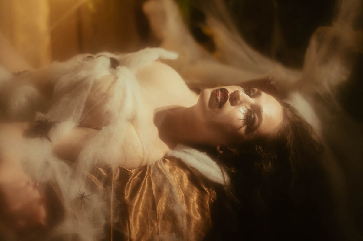A woman reclines on gold satin sheets wrapped in wispy cobweb netting with a small prop spider tucked nearby, her deep red lips parted and dark curls spread beneath her in warm golden light for this gothic glamour photoshoot at Royal Lune Photo.