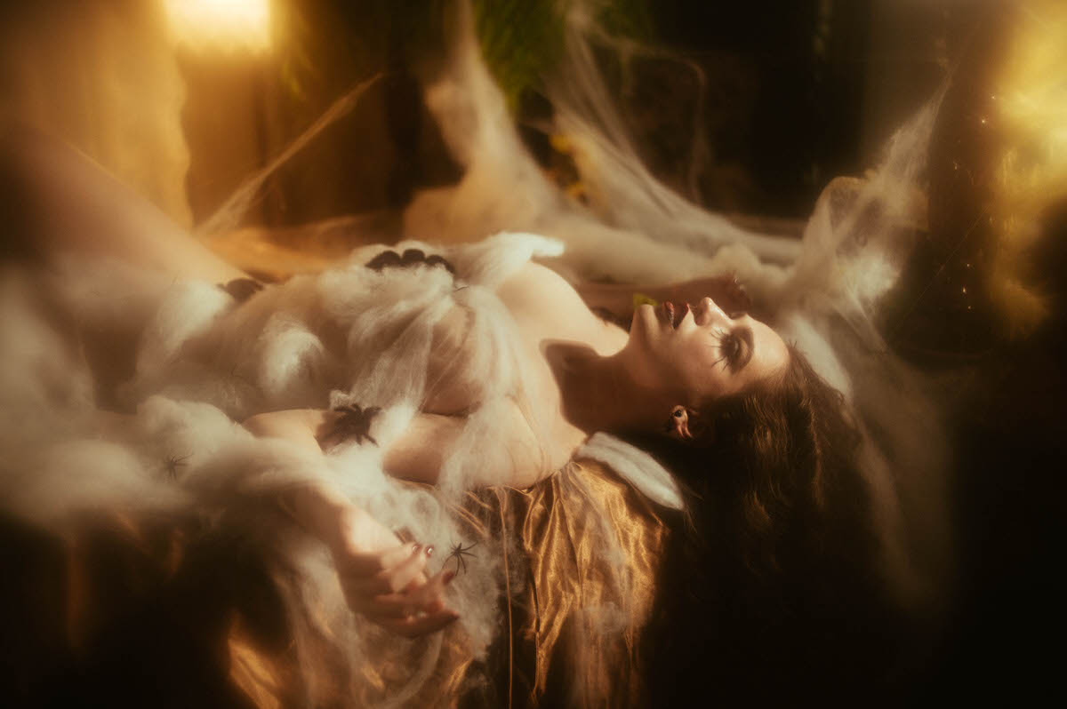 A woman lies back on gold satin sheets surrounded by sheer cobweb netting and small prop spiders, her spider leg lashes and dark red lips glowing in warm amber light for this sfx makeup photoshoot at Royal Lune Photo.