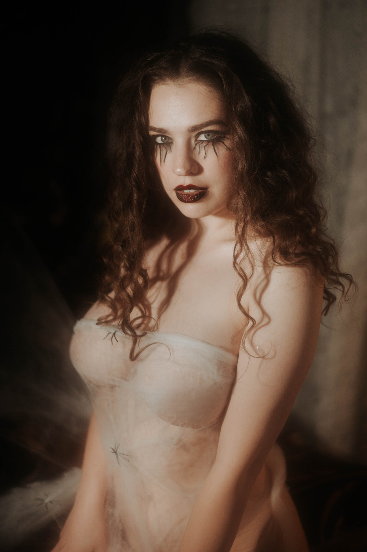 A woman poses in a sheer cobweb wrapped bandeau top with a small prop spider resting near her hip, deep red lips, and bold gothic eye makeup against a softly lit backdrop for this gothic boudoir photoshoot at Royal Lune Photo.