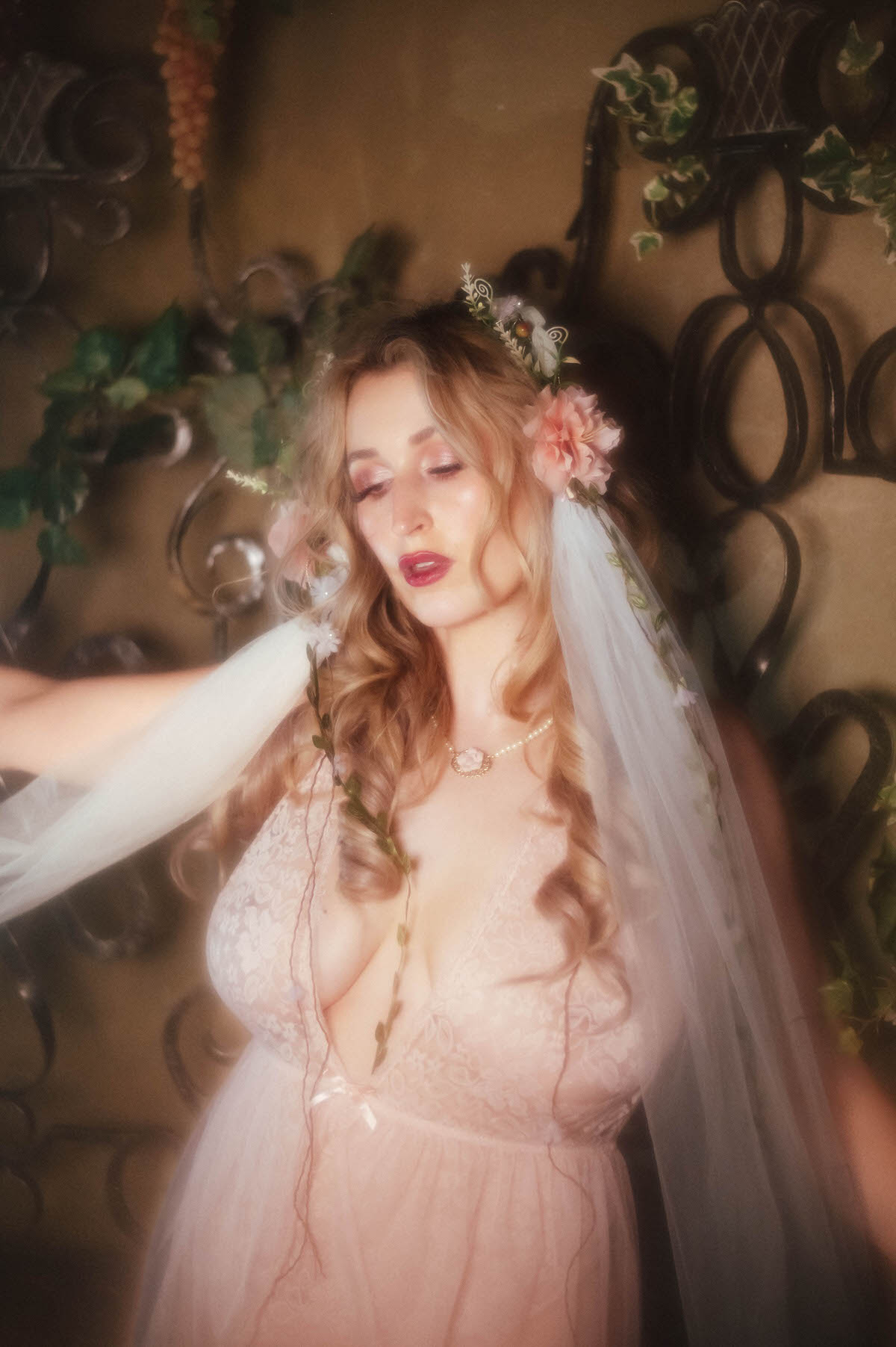 A close up shows a woman wearing a floral crown of blush and white blooms with a trailing veil and greenery draped down her chest, her vintage blush lace teddy visible beneath a pearl necklace against an ivy covered wrought iron backdrop, embodying the whimsical boudoir portraiture Dallas style of Royal Lune Photo.