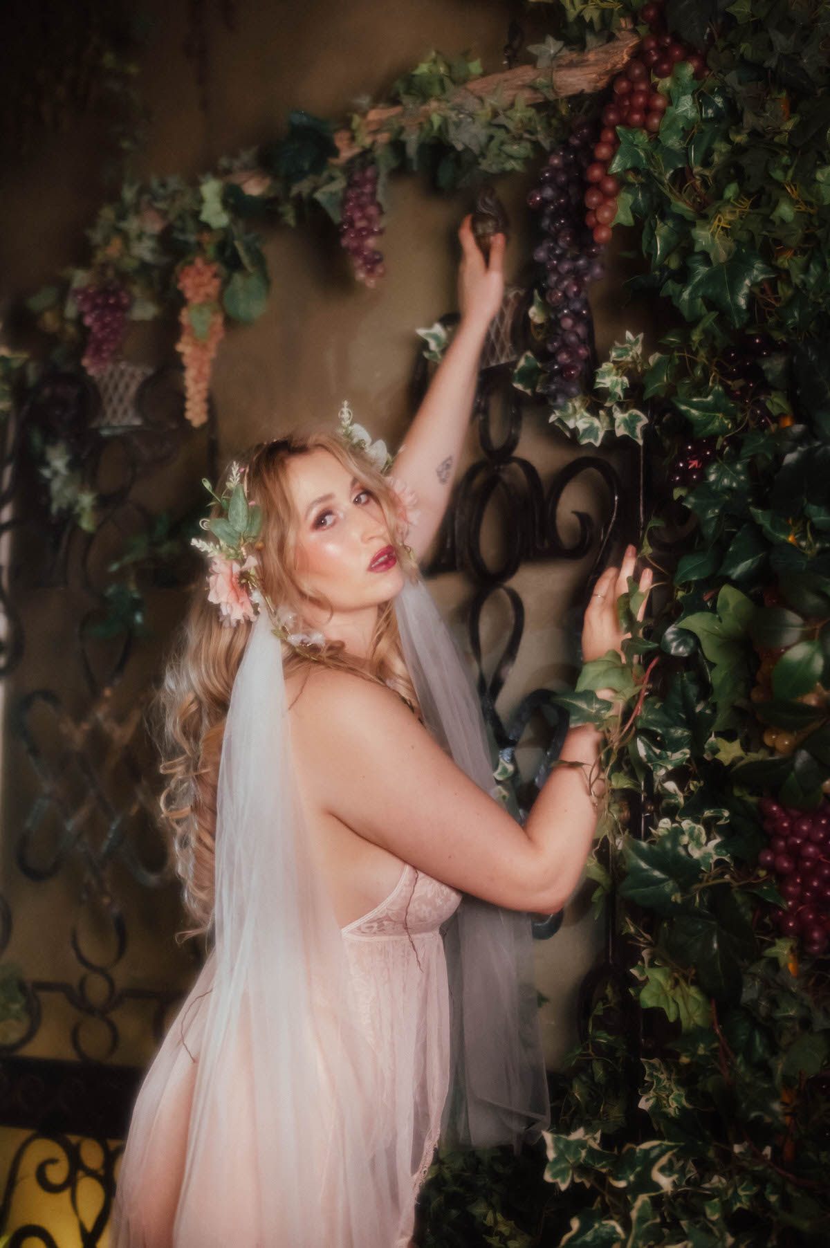 A woman glances over her shoulder while reaching toward ivy and grape clusters, her floral crown and trailing veil flowing down her back over a vintage blush lace teddy, capturing the soft light intimate portraiture of this dreamy boudoir photoshoot with Royal Lune Photo.