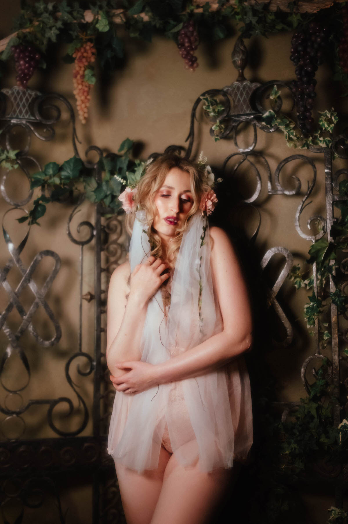 A woman stands with arms crossed against a wrought iron trellis draped in ivy and hanging grape clusters, wearing a vintage blush lace teddy and a floral crown with a trailing veil, capturing the Pre Raphaelite inspired finish of this dreamy boudoir photoshoot with Royal Lune Photo.