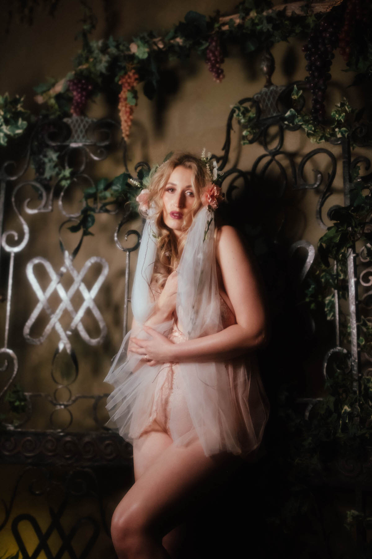 A woman clasps her hands at her chest with her veil pulled forward over her shoulders, wearing a vintage blush lace teddy with tulle detailing and a floral crown, standing before a wrought iron gate framed by ivy and grape clusters, part of this dreamy boudoir photoshoot with Royal Lune Photo.