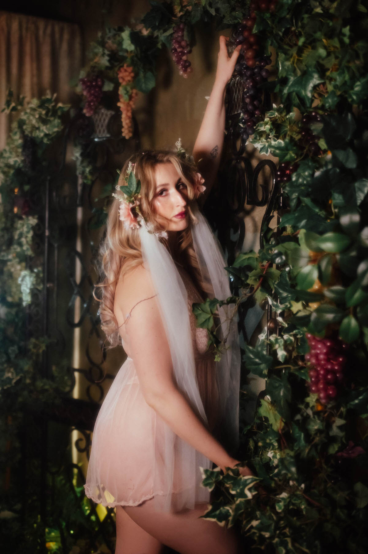 A woman in profile reaches up to touch hanging grape clusters, her trailing veil and vintage blush lace teddy catching the light against a backdrop of ivy and wrought iron, showcasing the fine art boudoir photography Dallas style of Royal Lune Photo.