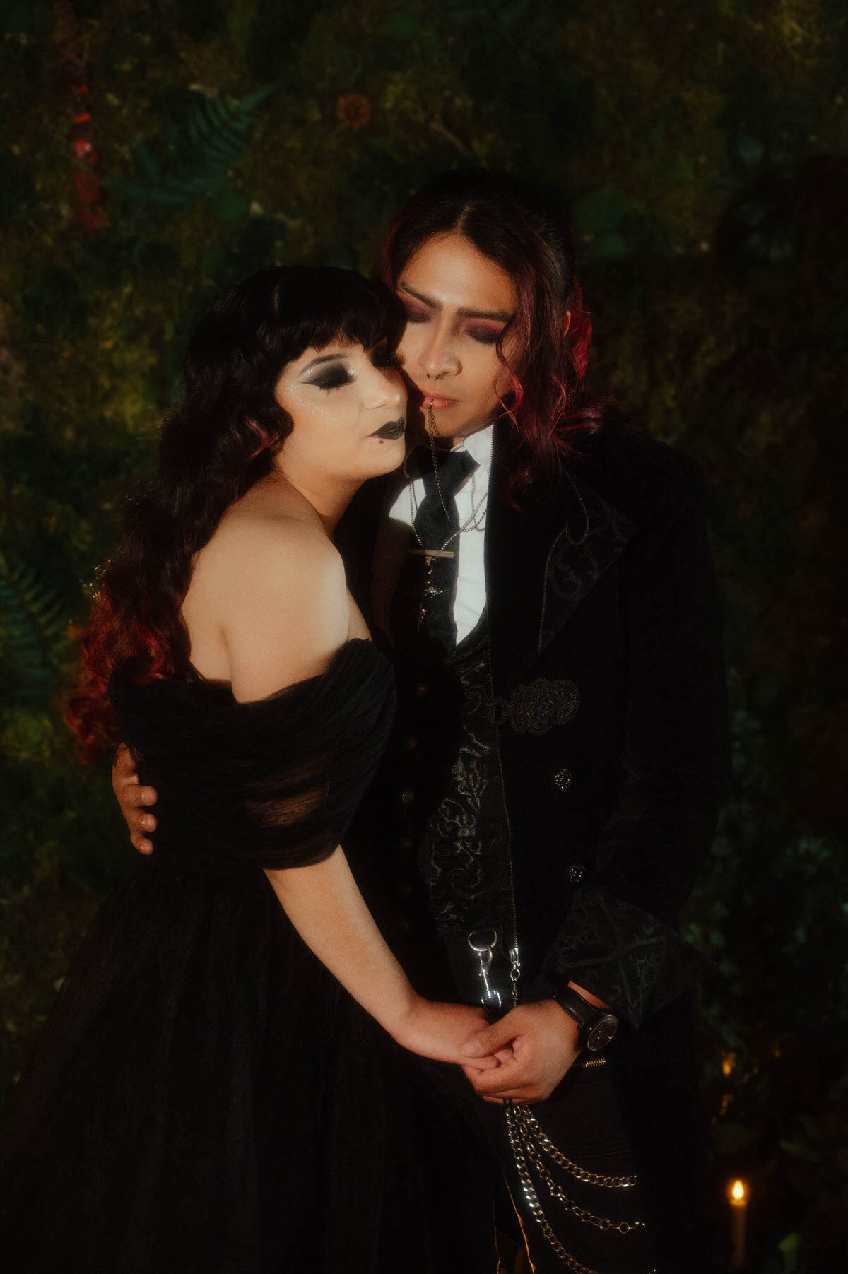 Couple in a black tulle gown and velvet coat holding hands against a leafy backdrop for gothic couples photography