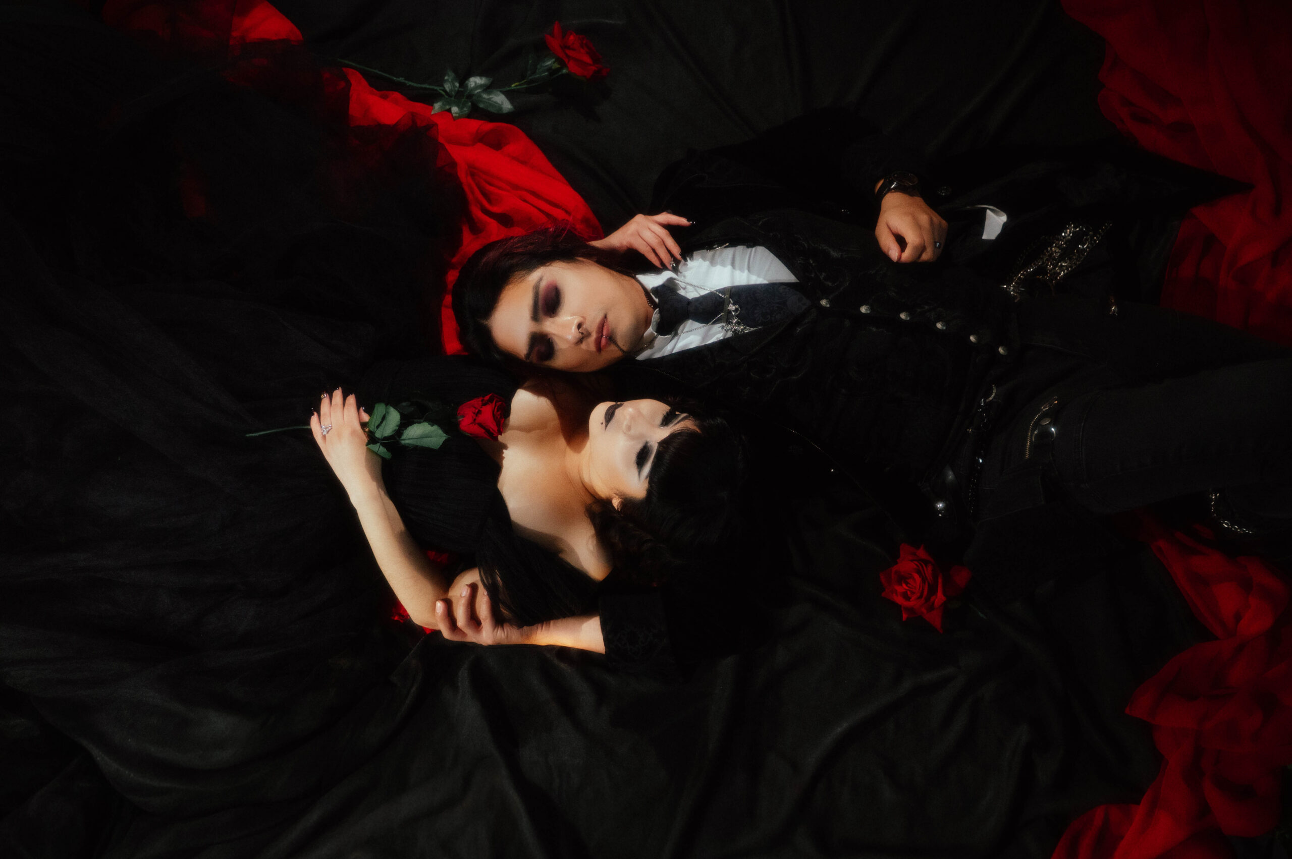 Couple in gothic outfits lying on black fabric with red roses, taken by an alternative engagement photographer in Dallas