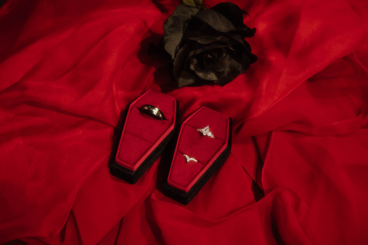 Wedding rings in coffin-shaped red velvet boxes on red fabric beside a black rose for gothic couples photography