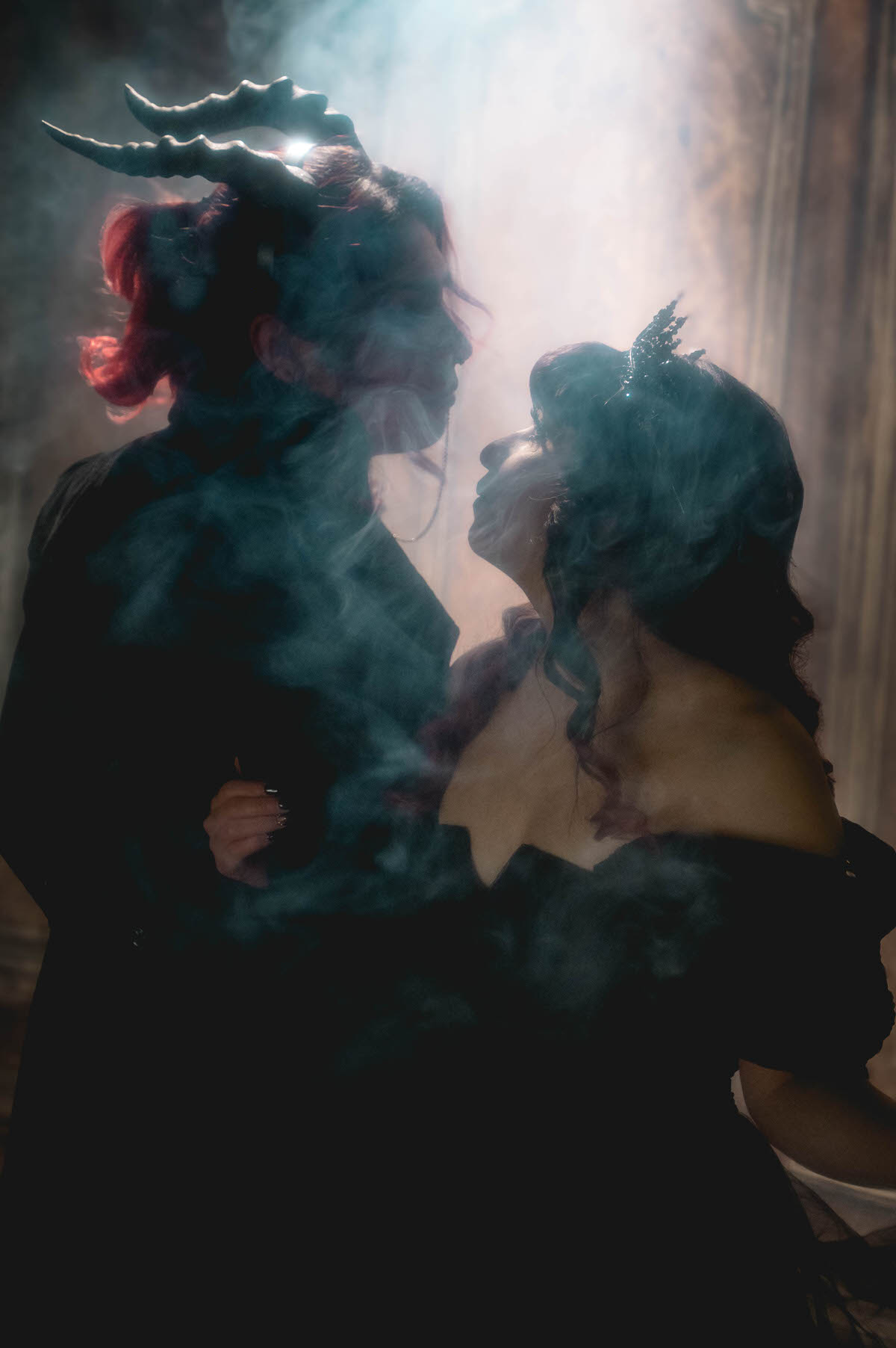 Couple in a horned headpiece and black crown facing each other in backlit fog for gothic engagement photography in Dallas