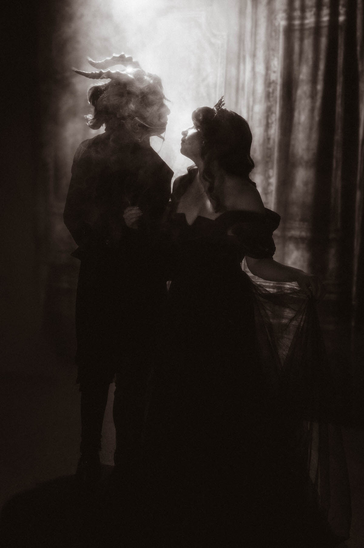 Black and white silhouette of a couple facing each other in backlit fog before curtains for dark fantasy engagement photos