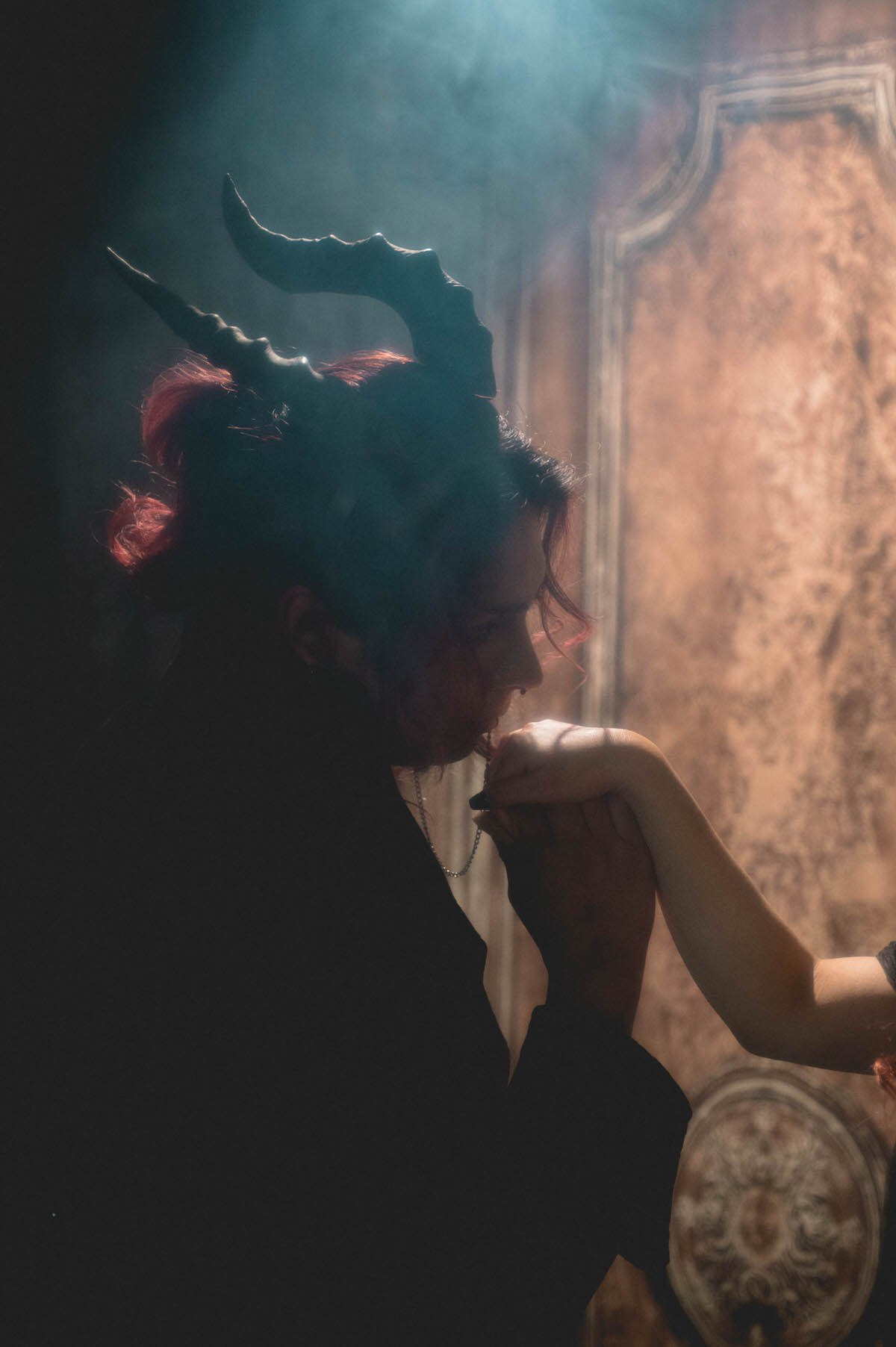 Silhouette of a person in a horned headpiece kissing a partner's hand in fog for dark romantic engagement photography